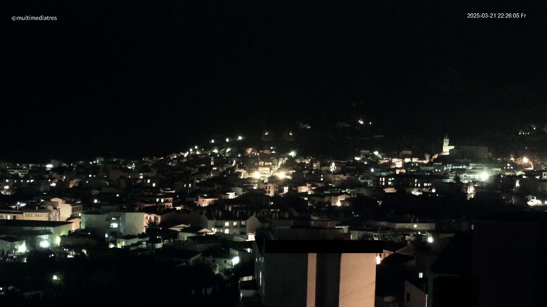 A nighttime view of a town nestled on a hillside, illuminated by scattered lights under a clear, dark sky.