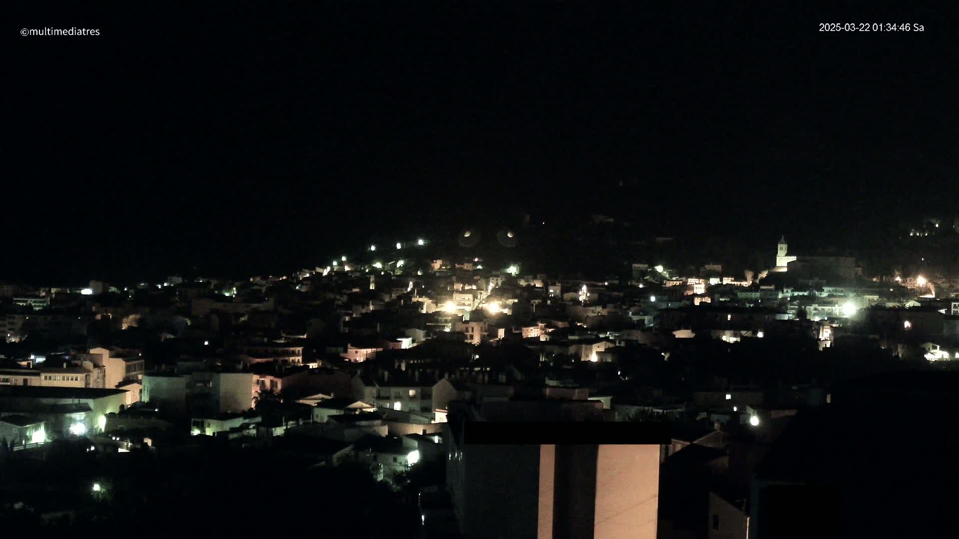 A nighttime long shot of a town nestled on a hillside, with scattered lights illuminating the buildings under a dark sky.