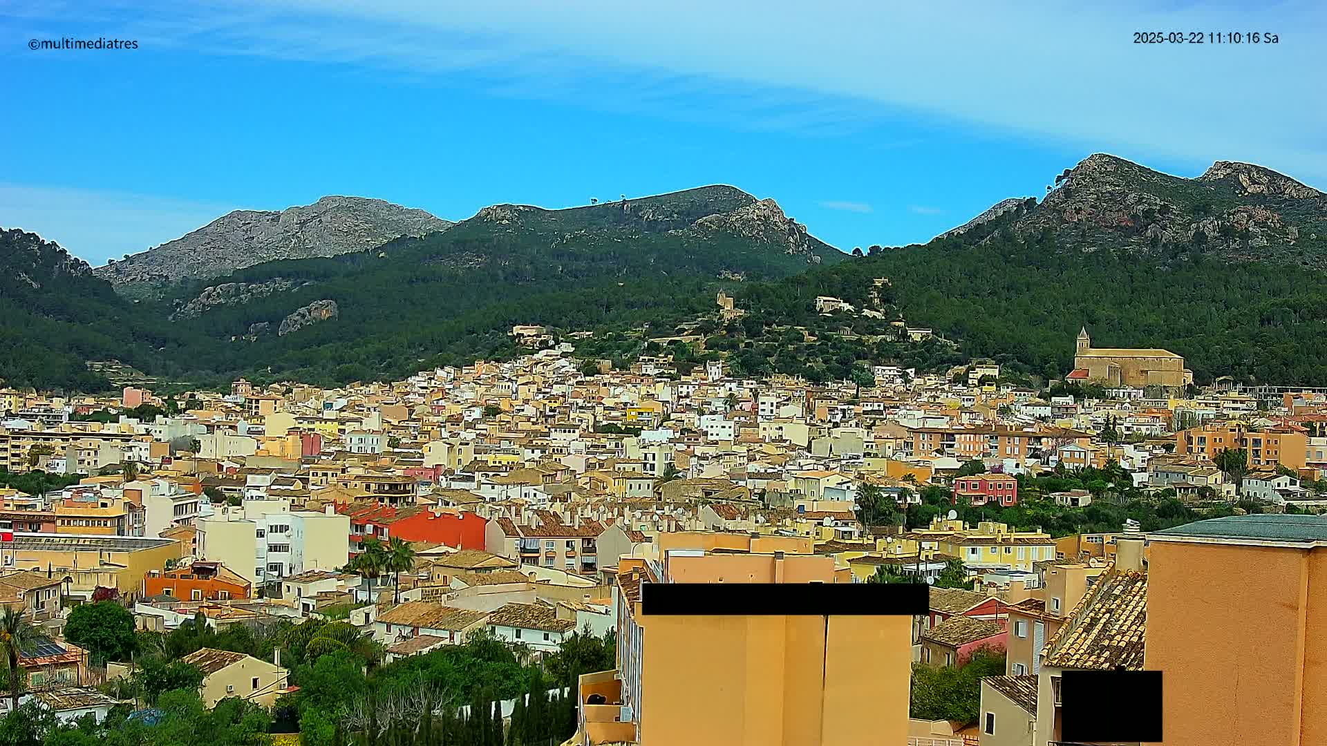 A sprawling town of low-rise buildings nestles in a valley surrounded by verdant, rocky mountains under a clear blue sky.