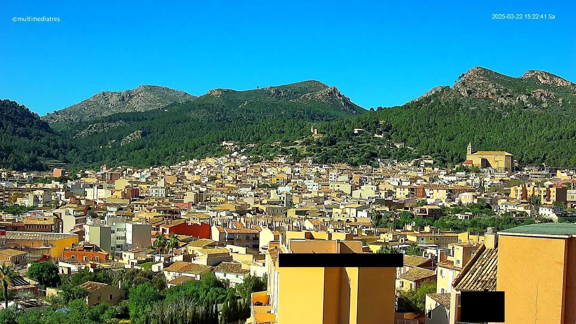 A dense collection of low-rise buildings stretches across a hillside, nestled beneath verdant mountains under a clear blue sky.