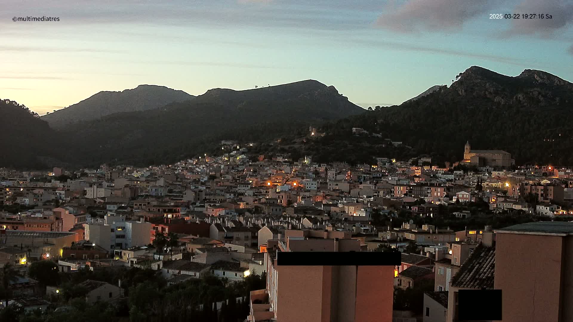 A town with many lights nestled at the base of dark, mountainous terrain under a twilight sky.