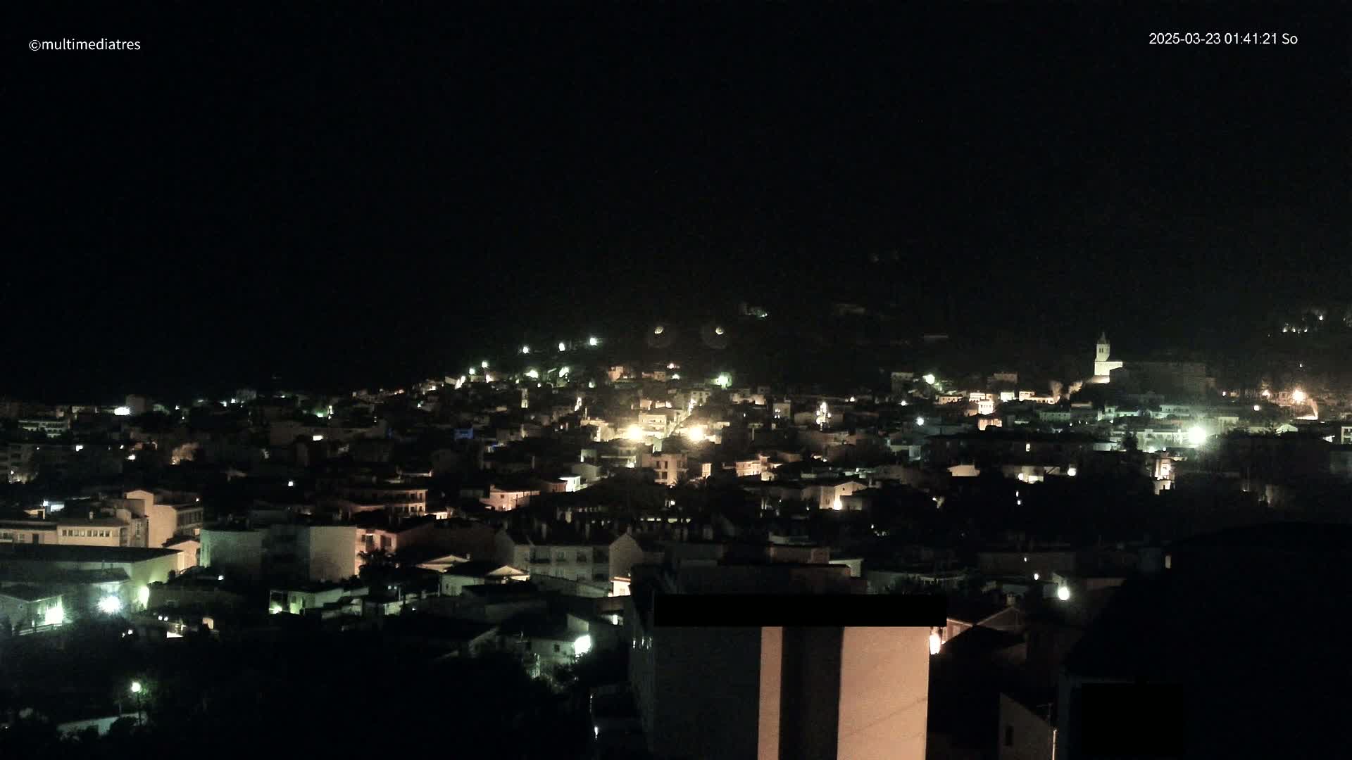 A nighttime, aerial view of a town nestled on a hillside, illuminated by scattered lights.
