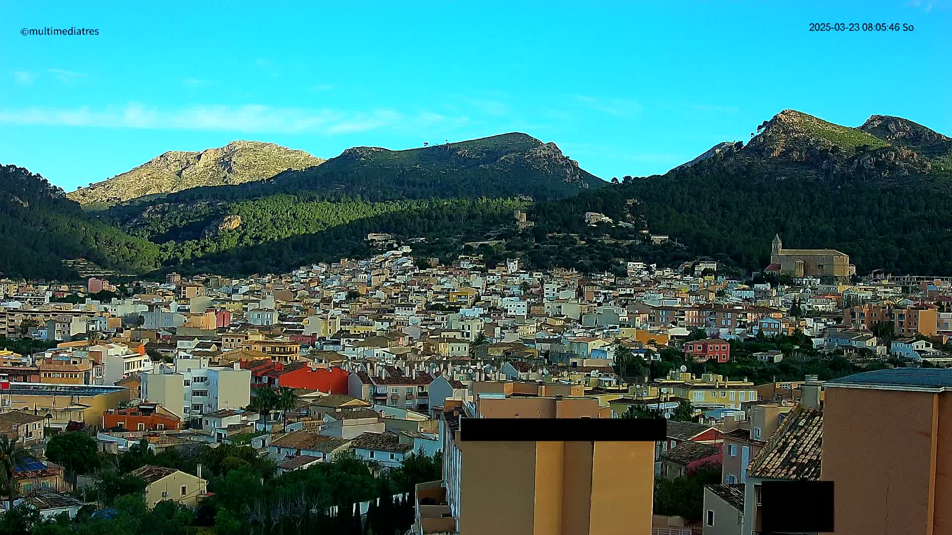 A sprawling town of low-rise buildings nestled at the base of several verdant, rocky mountains under a clear blue sky.