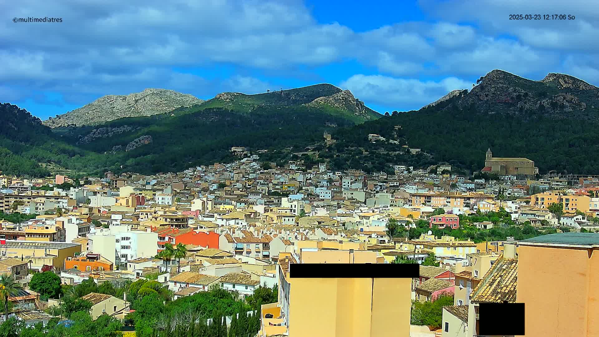 A sprawling town of low-rise buildings nestles at the base of several large, green-covered mountains under a partly cloudy sky.