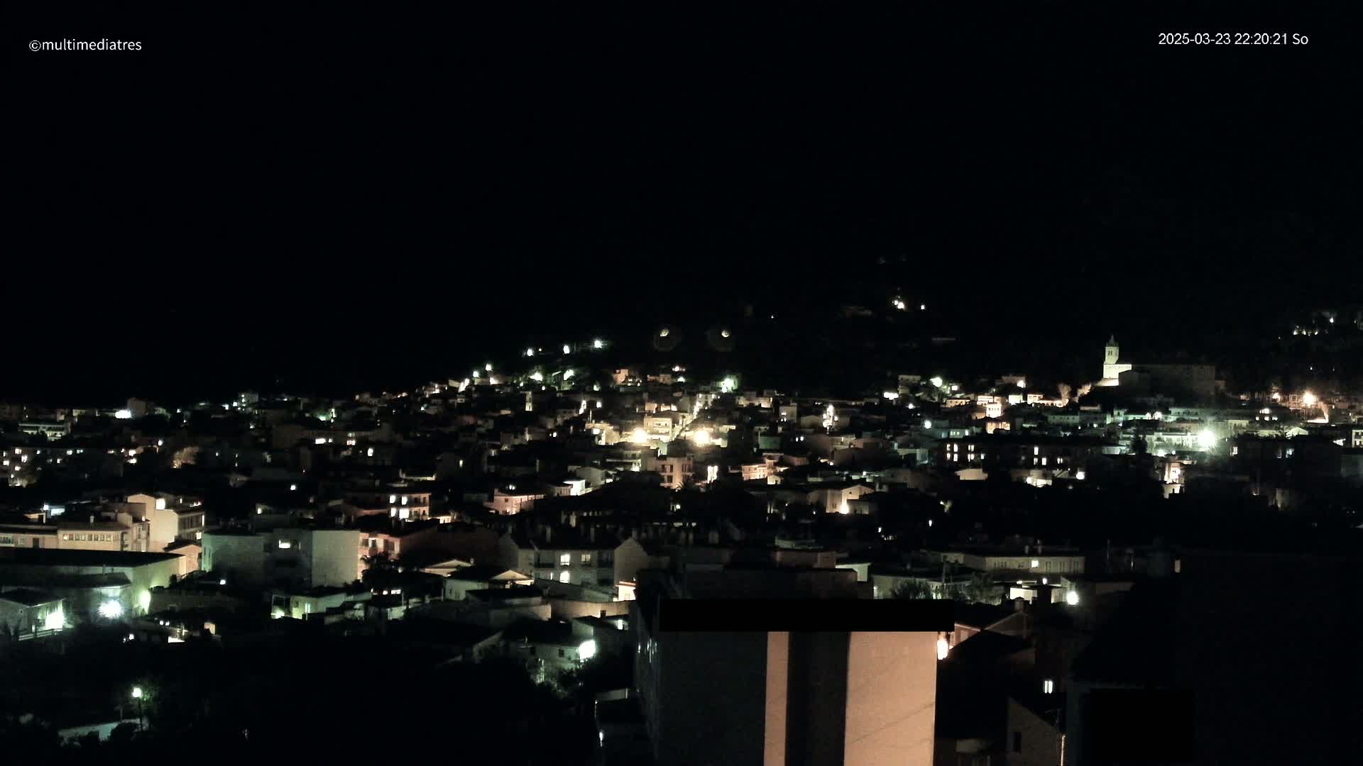 A nighttime view of a town nestled on a hillside, illuminated by the lights of numerous buildings.