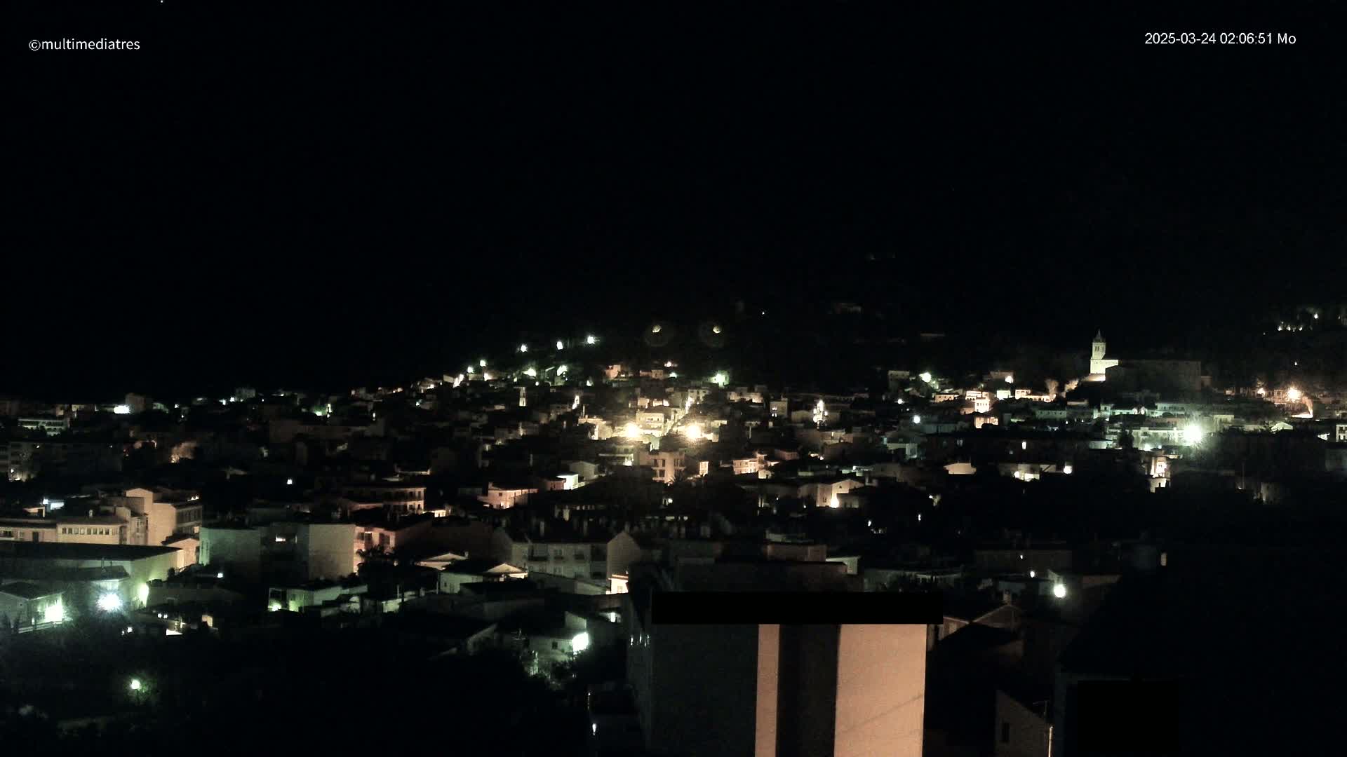 A nighttime view of a town nestled on a hillside, illuminated by scattered lights under a clear, dark sky.
