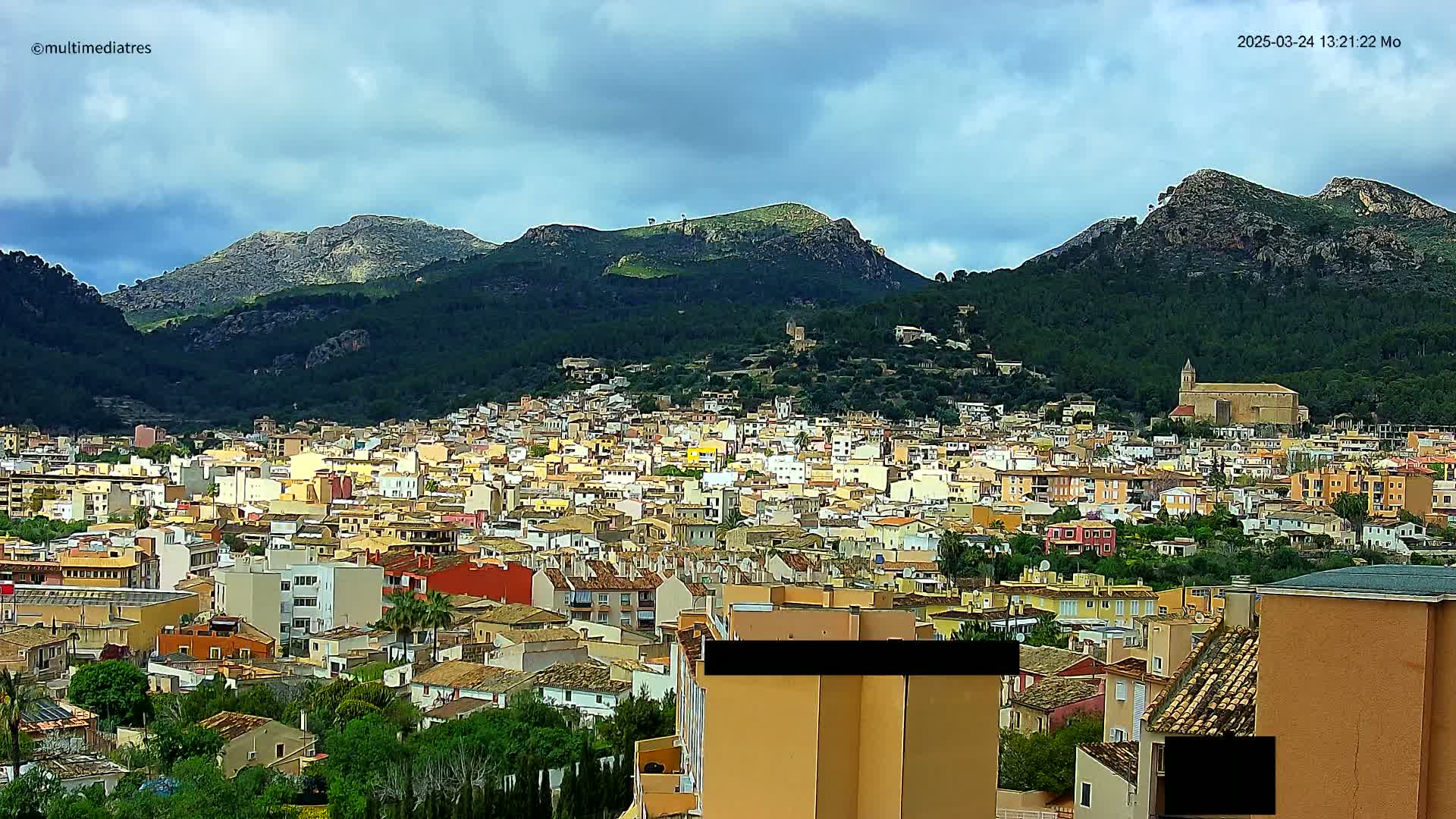 A sprawling town of tan and red-roofed buildings nestled at the foot of verdant, rocky mountains under a partly cloudy sky.