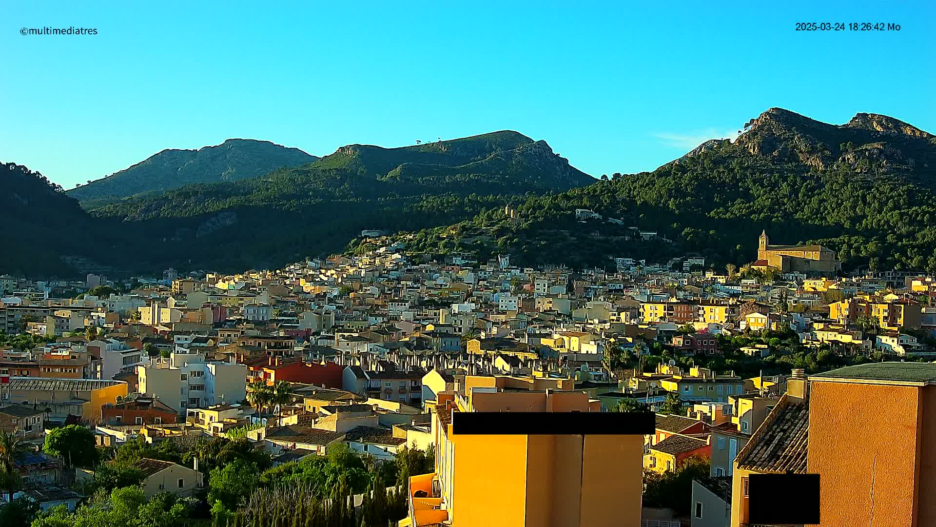 A sprawling town of low-rise buildings nestles at the base of lush, green mountains under a clear, blue sky.