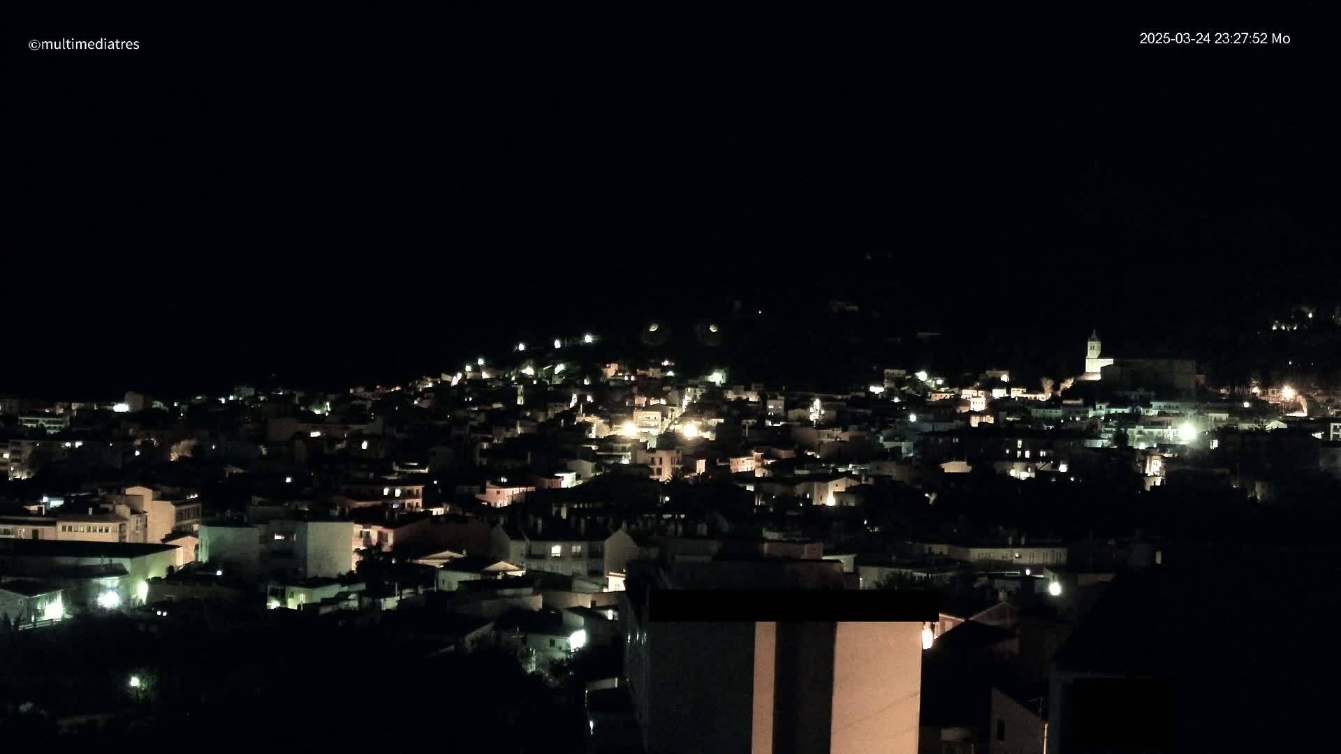 A nighttime view of a town nestled on a hillside, with scattered lights illuminating buildings under a dark sky.