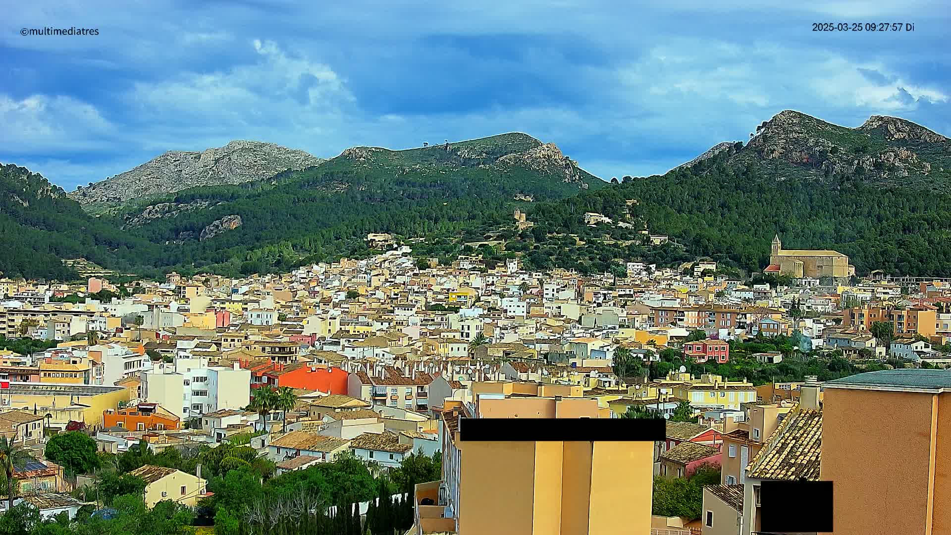 A sprawling town of colorful buildings nestles at the base of verdant, rocky mountains under a partly cloudy sky.