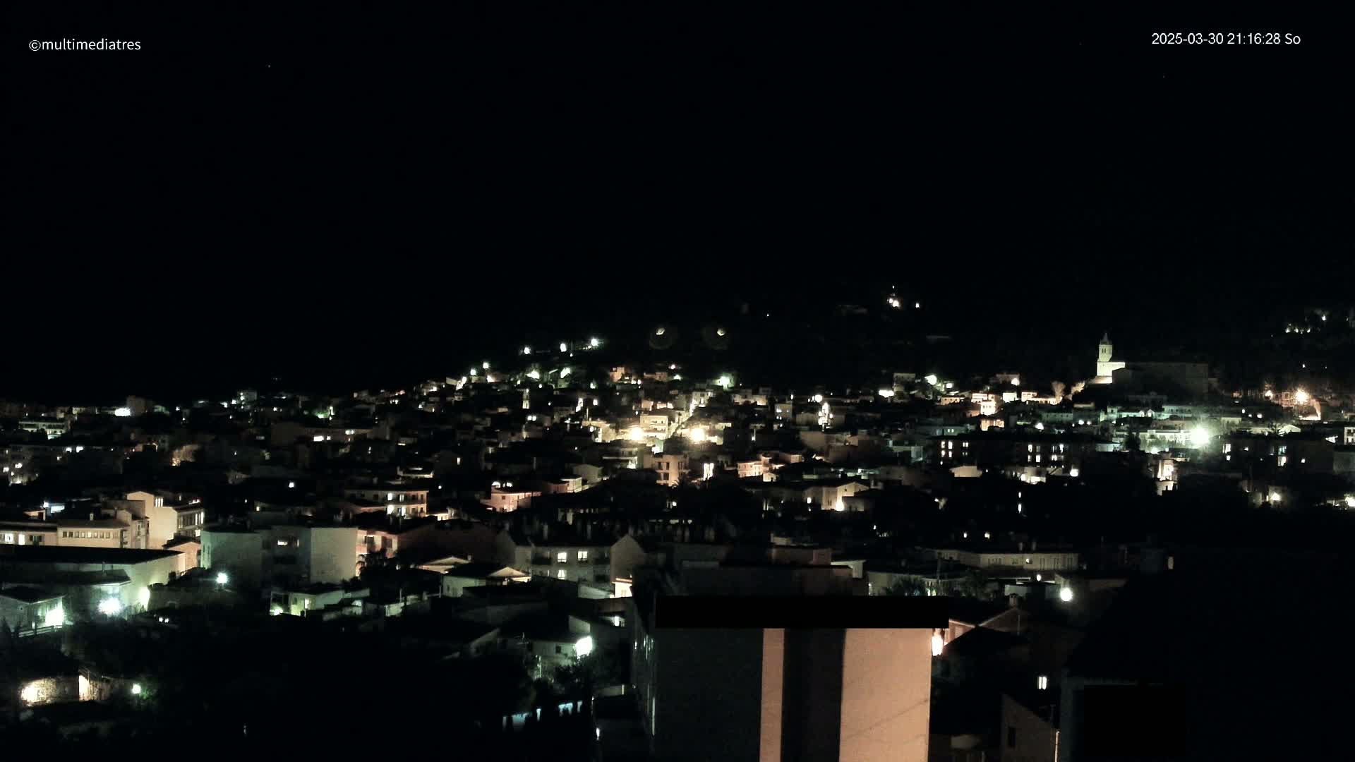A nighttime view of a town nestled on a hill, illuminated by scattered lights under a clear, dark sky.