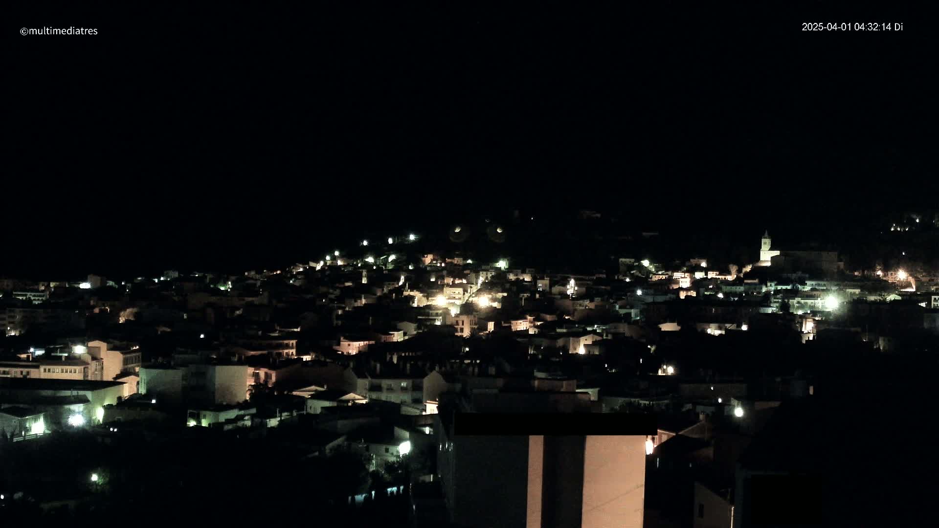 A nighttime, aerial view of a town nestled on a hillside, illuminated by scattered lights.