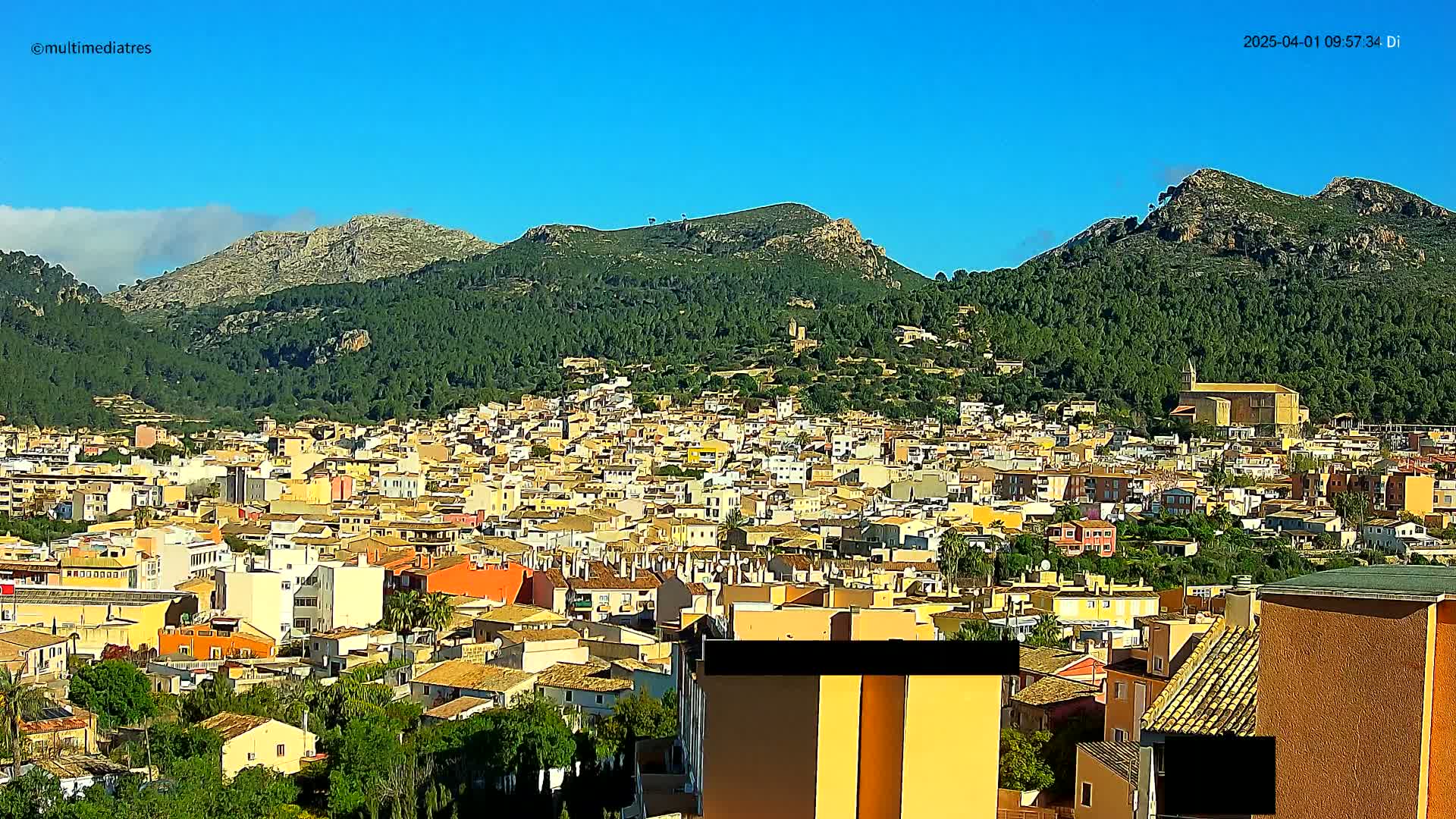 A sprawling town of low, light-colored buildings nestled at the base of verdant, rocky mountains under a clear blue sky.