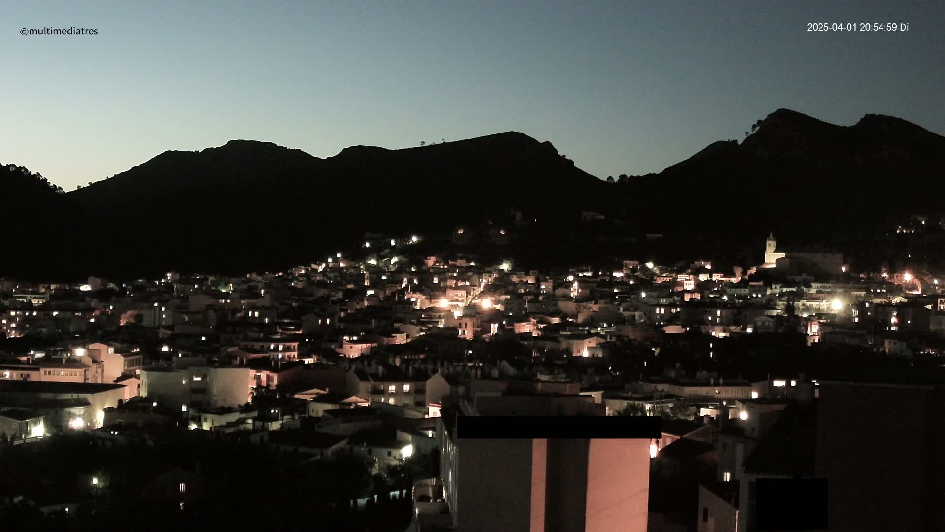 A nighttime view of a town nestled in a valley, illuminated by scattered lights, with dark mountains silhouetted against a twilight sky.