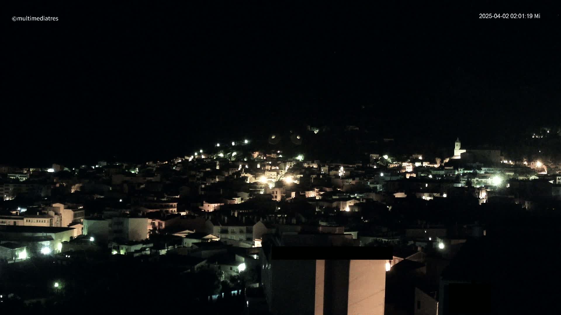 A nighttime, high-angle view of a town nestled on a hillside, with the lights of numerous buildings visible against the dark sky.