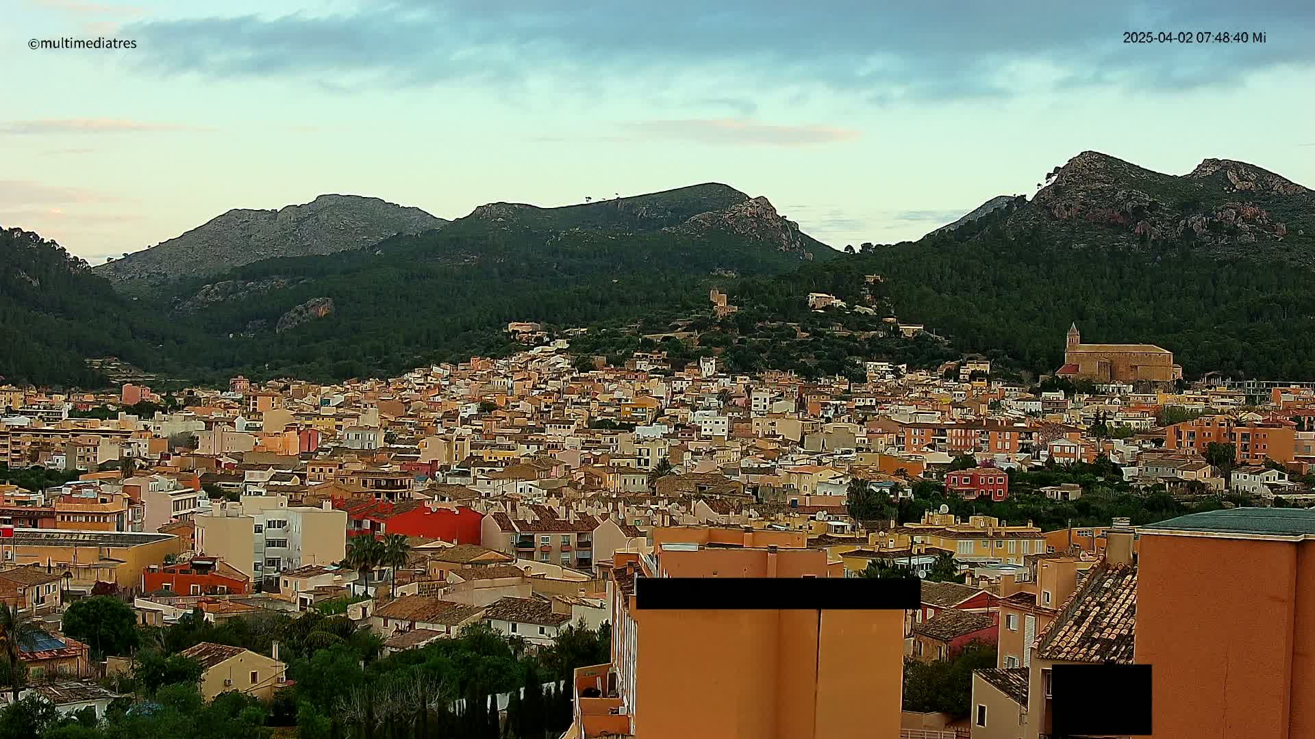 A sprawling town of light brown and tan buildings nestles in a valley, backed by dark green hills under a mostly clear, dusky sky.