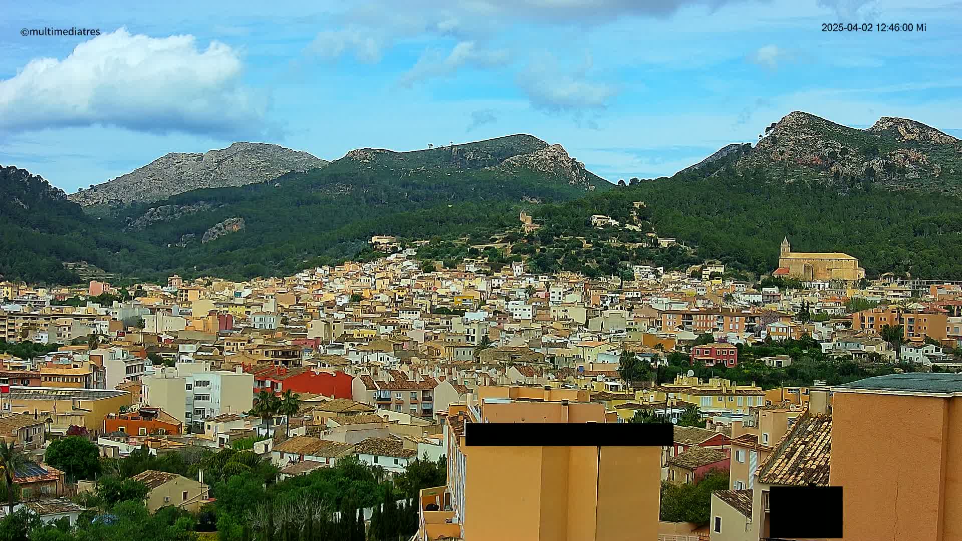 A sprawling town of pastel-colored buildings nestled in a valley, surrounded by lush green hills and mountains under a partly cloudy sky.