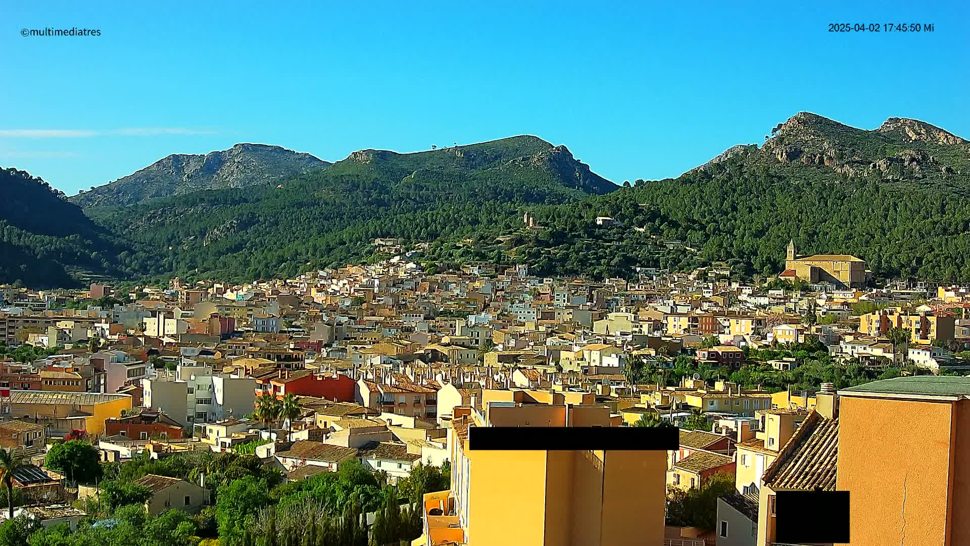 A sprawling town of low-rise buildings nestles at the foot of a mountain range under a clear blue sky.
