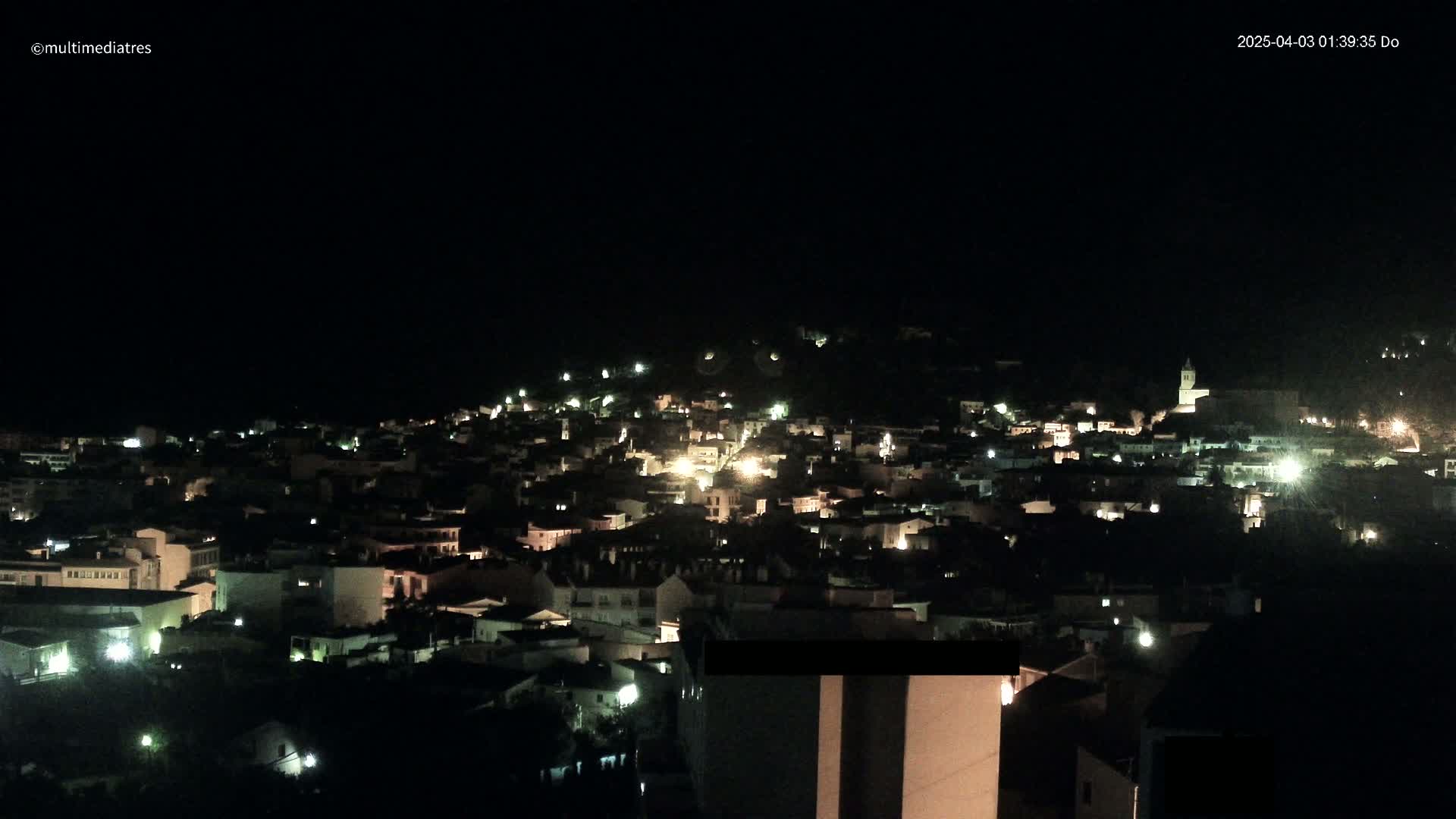 A nighttime, high-angle view of a town nestled on a hillside, with numerous lights illuminating the buildings and streets.