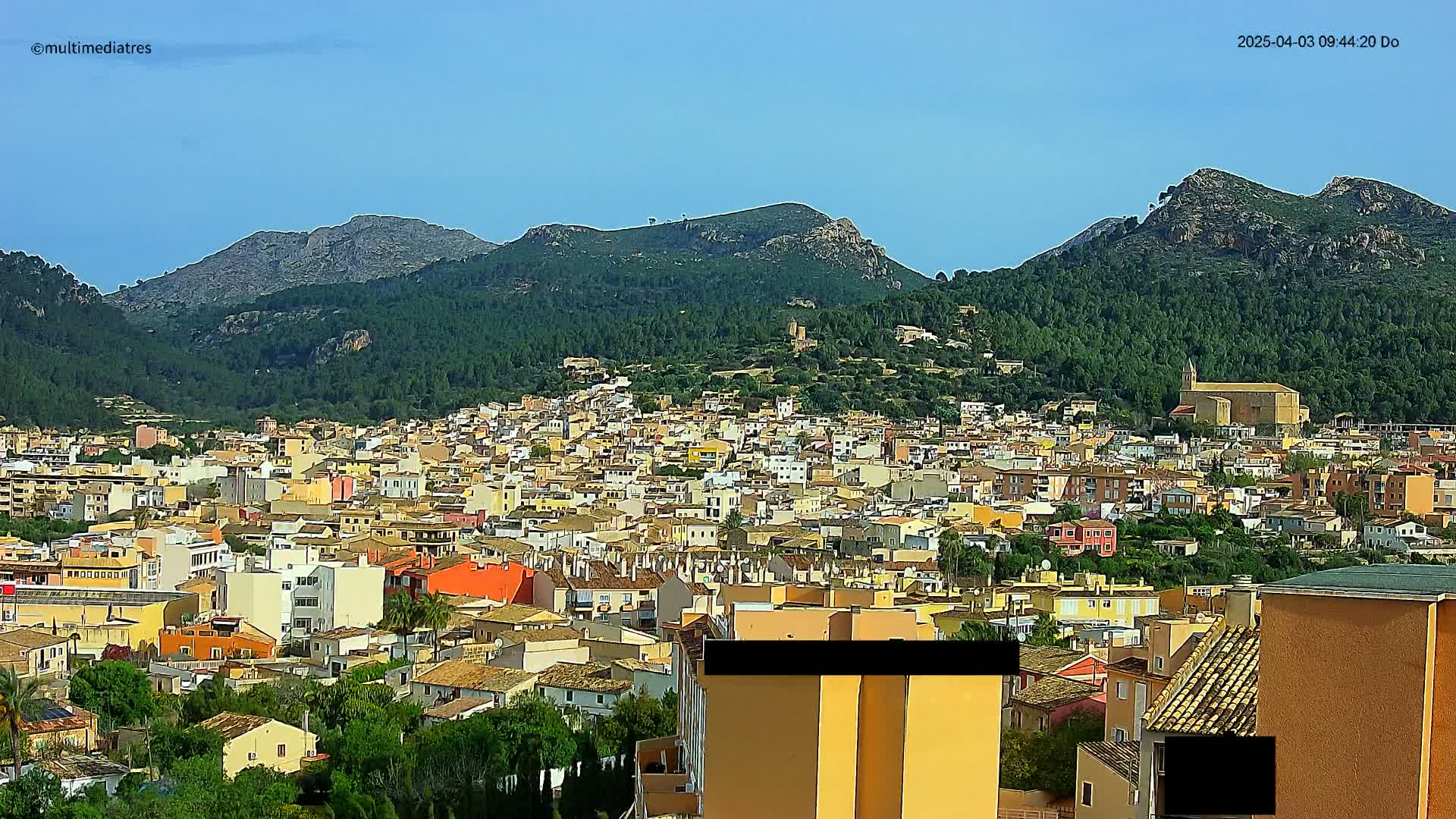 A sprawling town of light-colored buildings nestled against a backdrop of verdant, mountainous terrain under a clear blue sky.