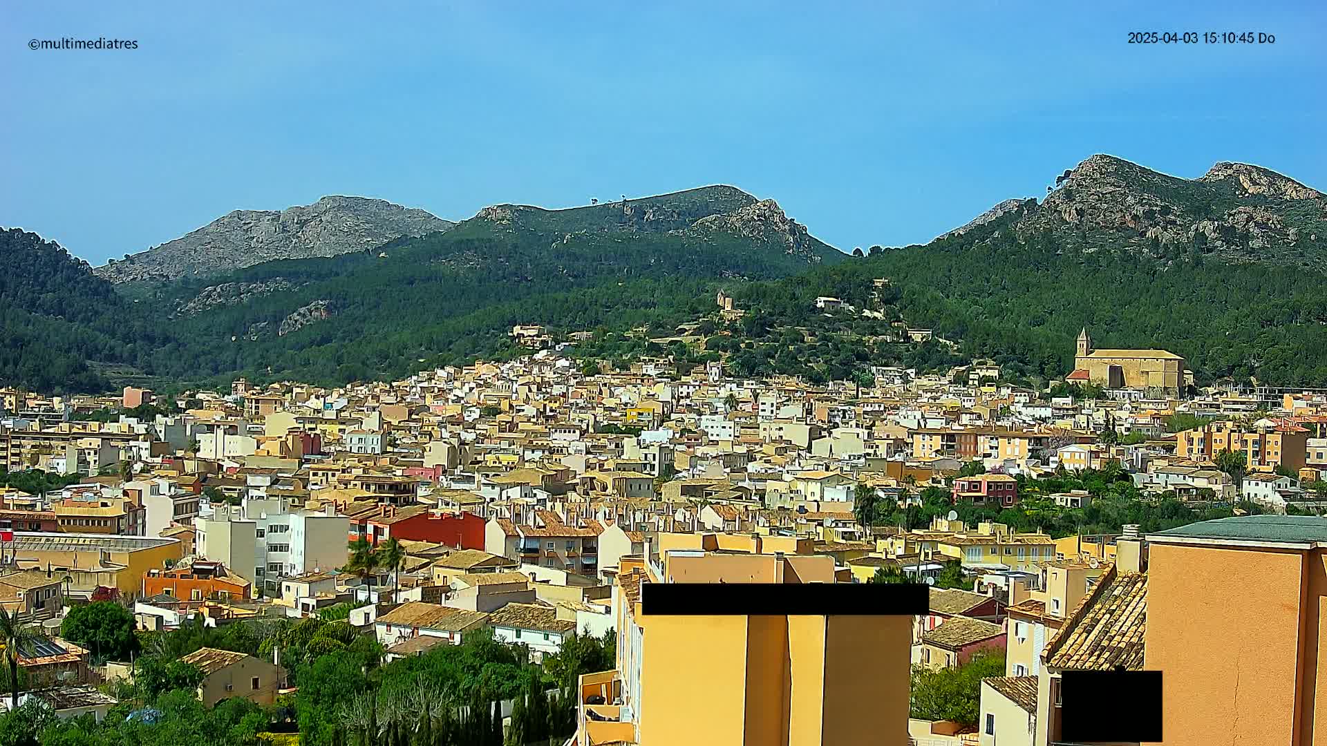 A sprawling town of tan and red-roofed buildings nestles at the foot of verdant, rocky mountains under a clear blue sky.