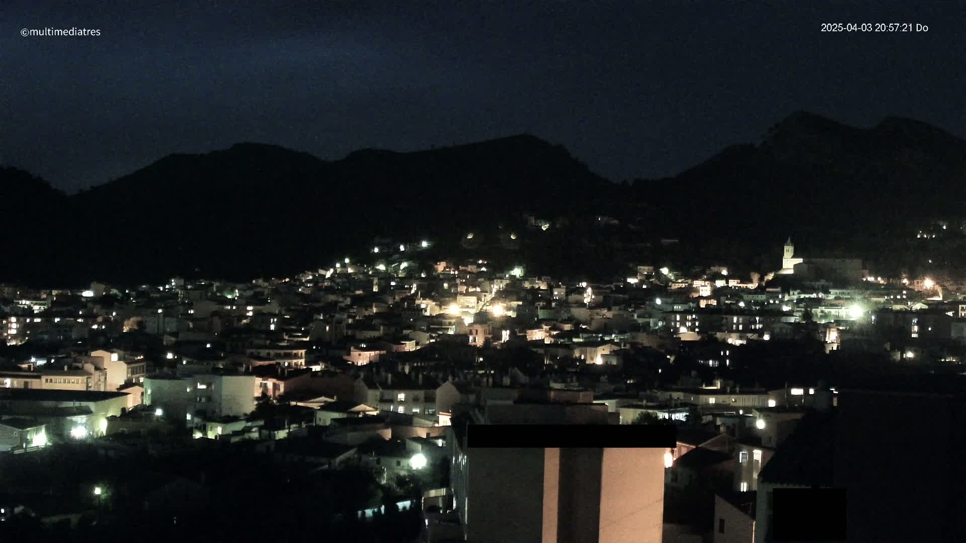 A nighttime view of a town nestled in a valley, illuminated by scattered lights, with dark mountains in the background under a clear, dark sky.