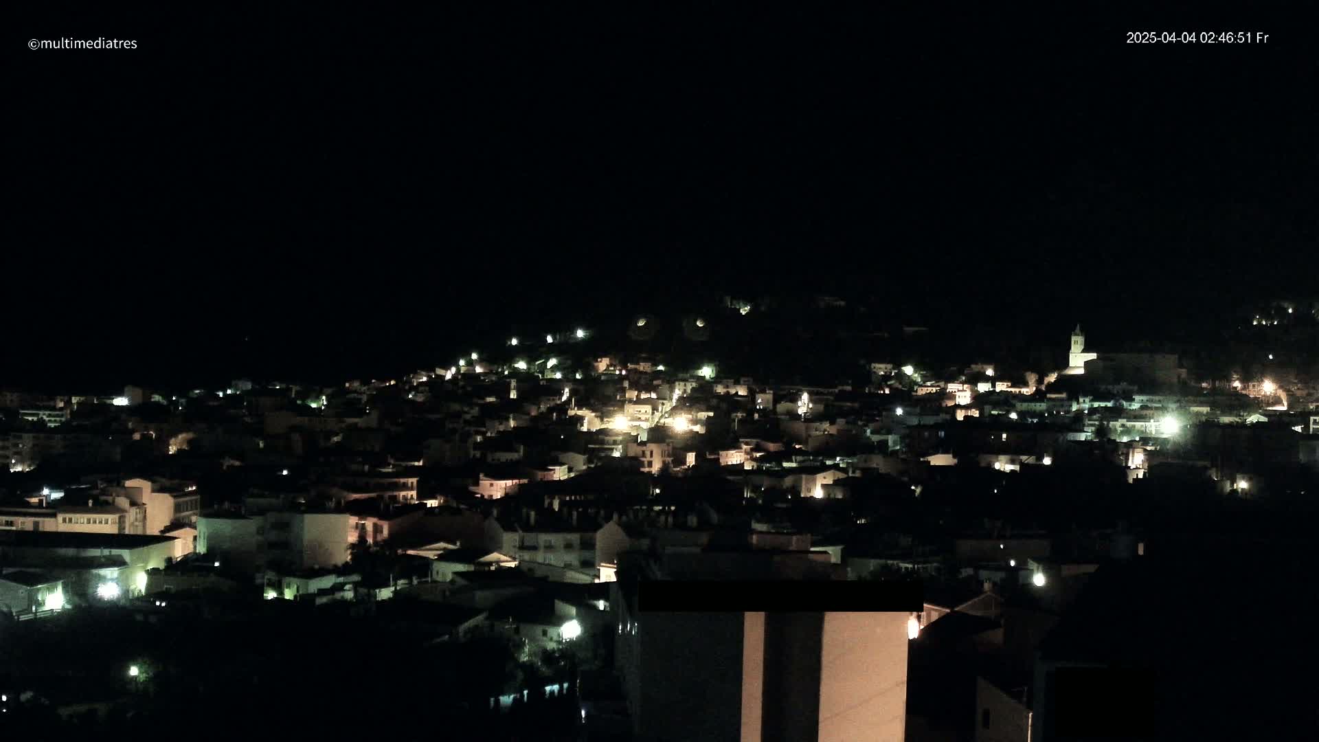 A nighttime, aerial view of a town's illuminated buildings spread across a hillside.