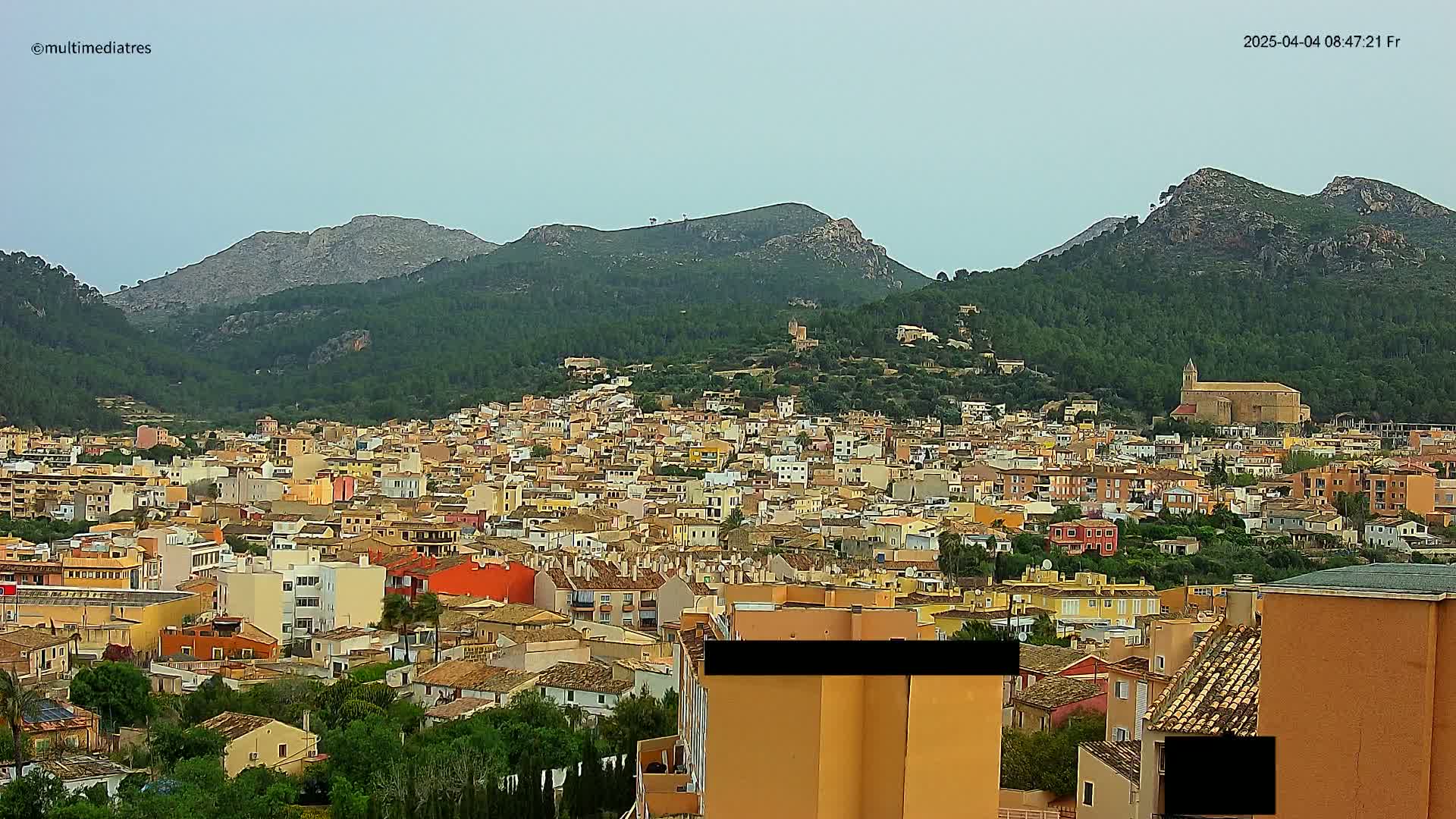 A sprawling town of tan and orange buildings nestles in a valley surrounded by verdant, mountainous terrain under a mostly clear sky.