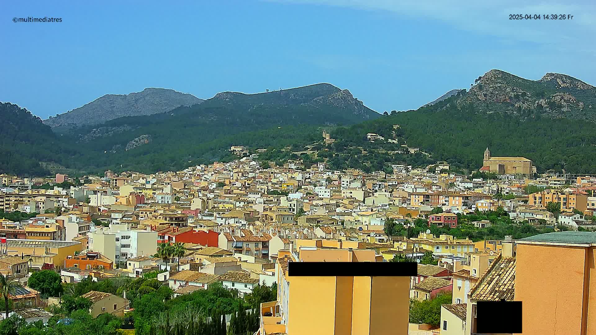 A sprawling town of low-rise buildings nestled in a valley, surrounded by lush green hills and mountains under a clear blue sky.
