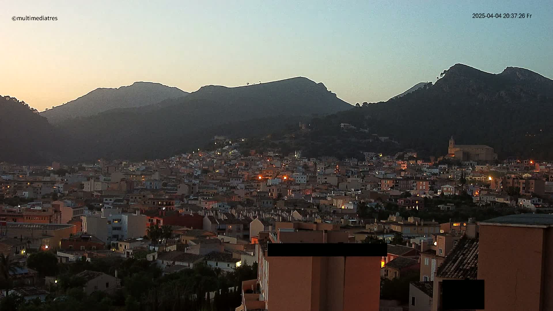A town nestled in a valley at dusk, illuminated by scattered lights, is overlooked by dark, mountainous terrain under a twilight sky.