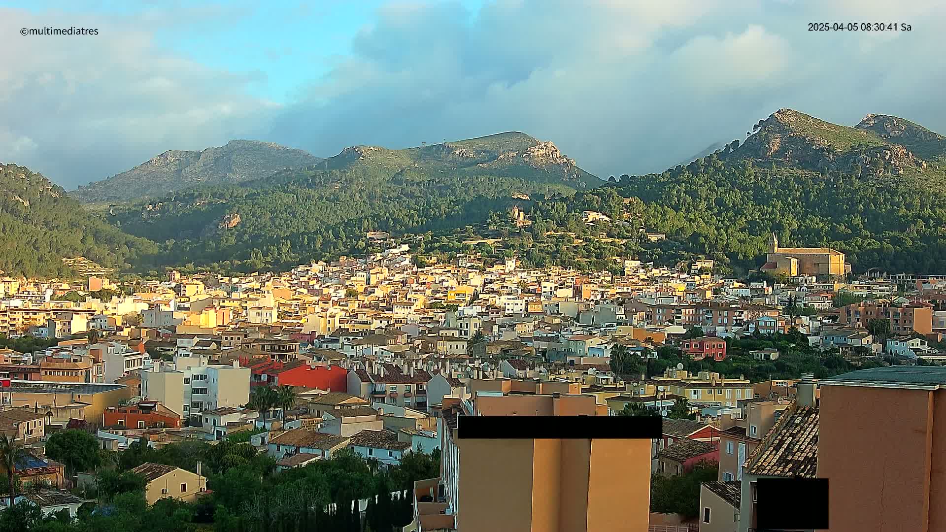 A sprawling town of light-colored buildings nestled in a valley, surrounded by lush green hills and mountains under a partly cloudy sky.