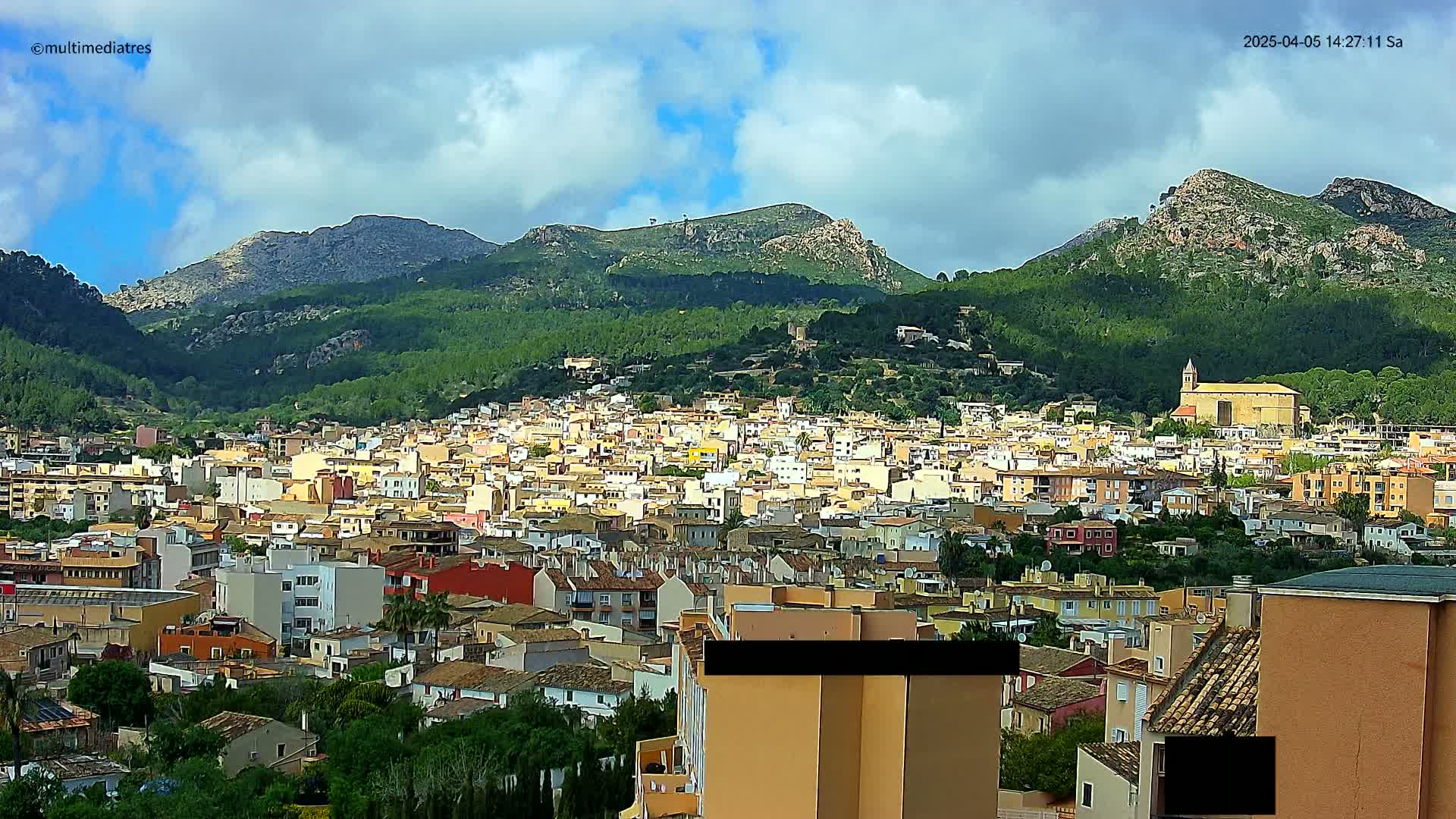 A sprawling town of light-colored buildings nestled in a valley, surrounded by lush green hills and mountains under a partly cloudy sky.