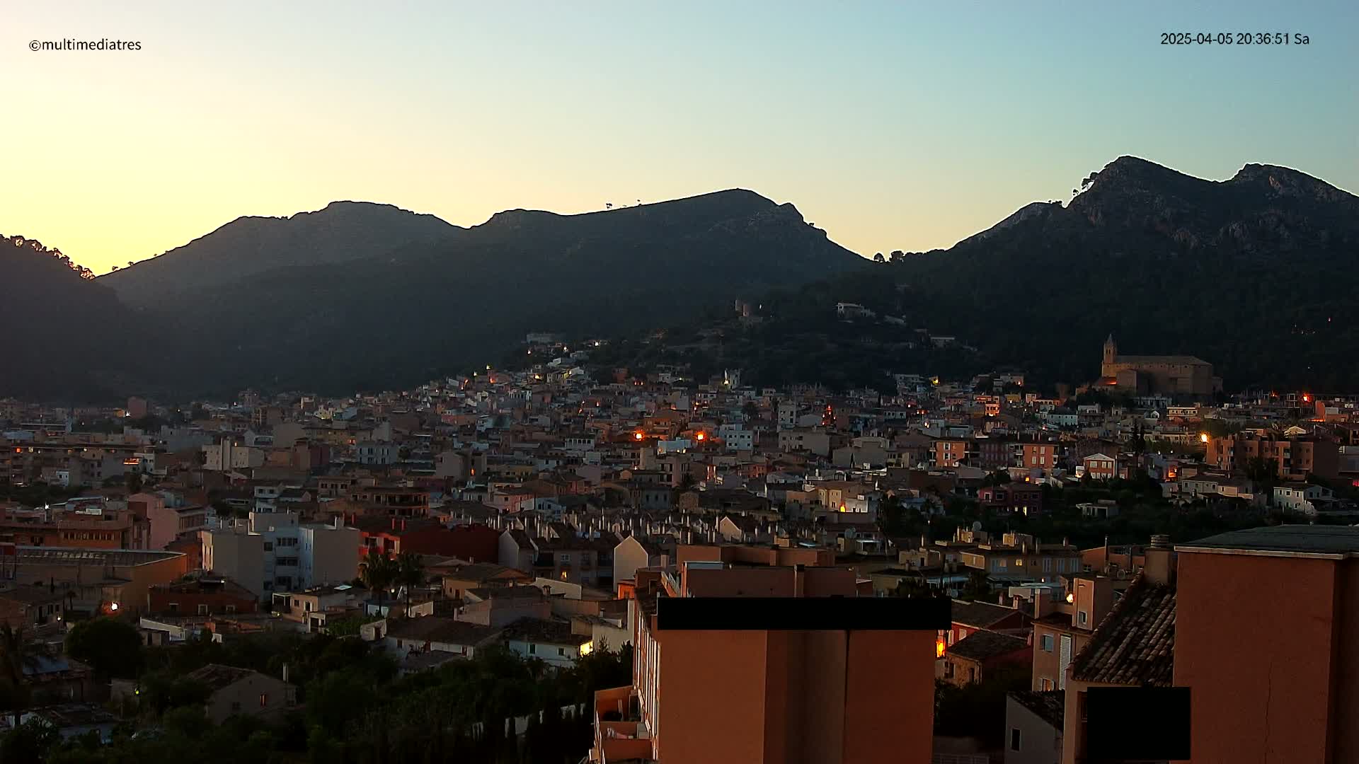 A sprawling town nestled at the base of dark, mountainous terrain during twilight.