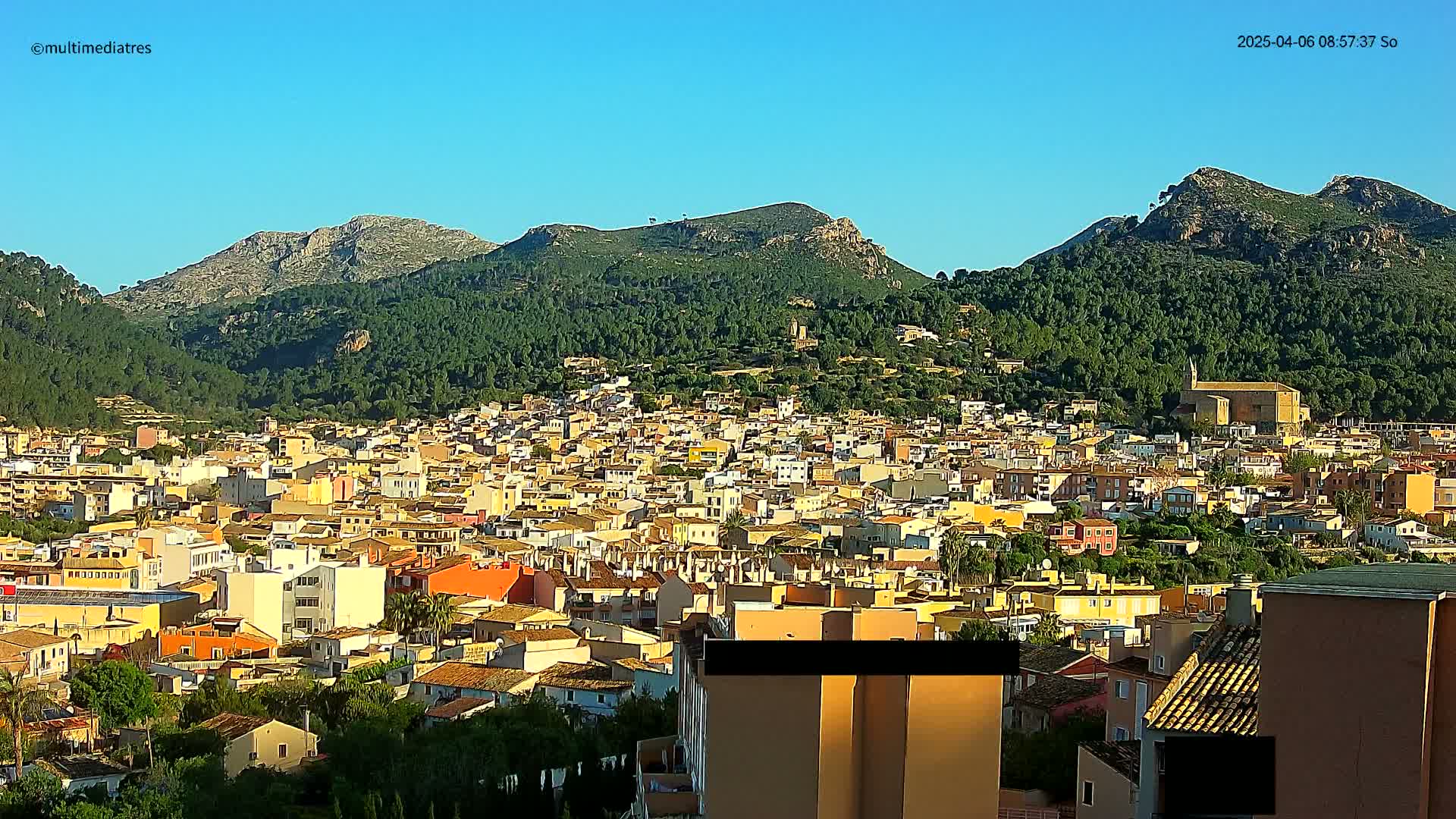 A dense collection of low-rise buildings situated at the base of a large, lush green mountain under a clear blue sky.