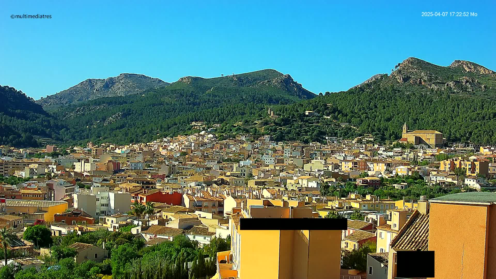 A sprawling town of low-rise buildings nestled in a valley surrounded by lush green hills under a clear blue sky.