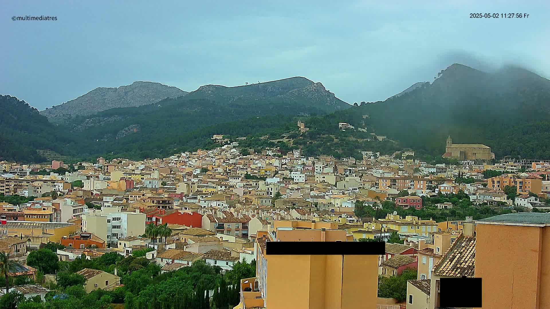 A sprawling town of low-rise buildings nestled in a valley between mountains, under a cloudy sky.