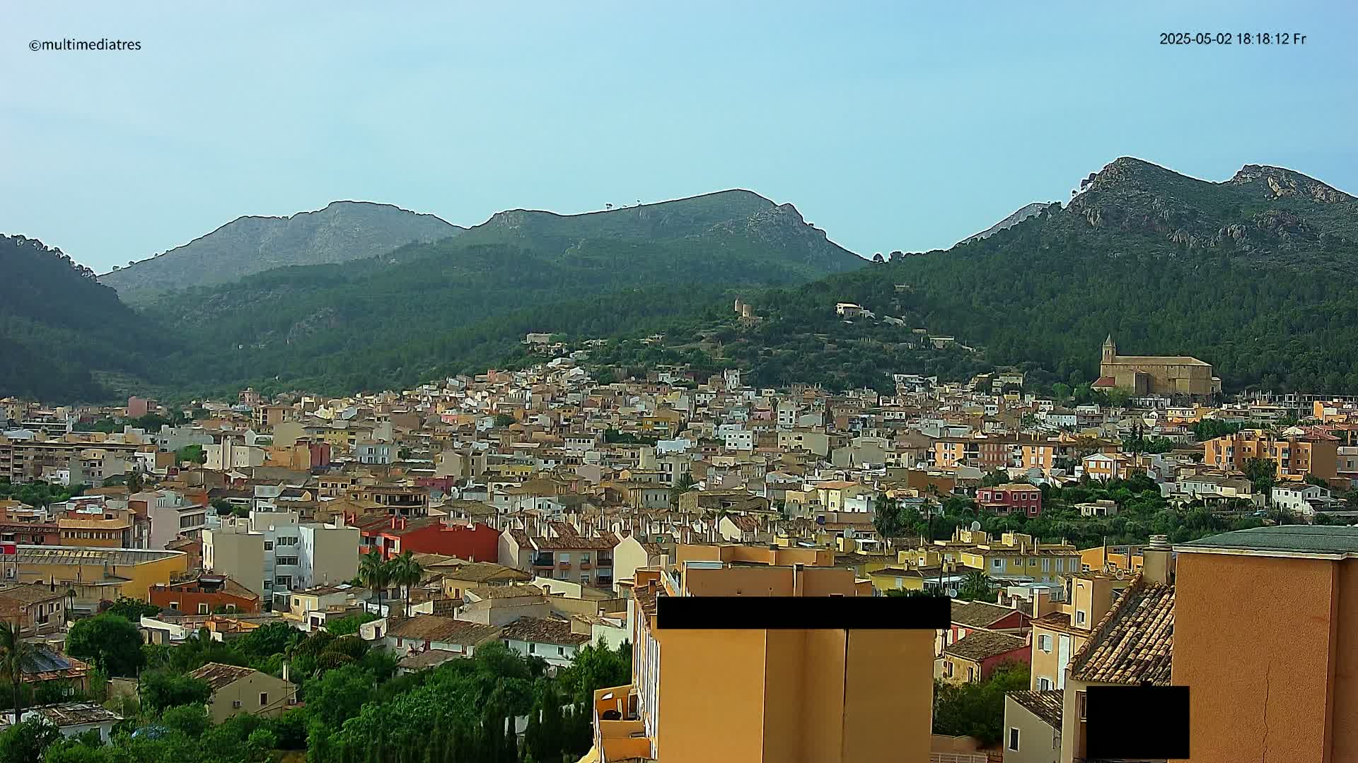 A sprawling town of low-rise buildings nestles at the foot of verdant, mountainous terrain under a clear sky.