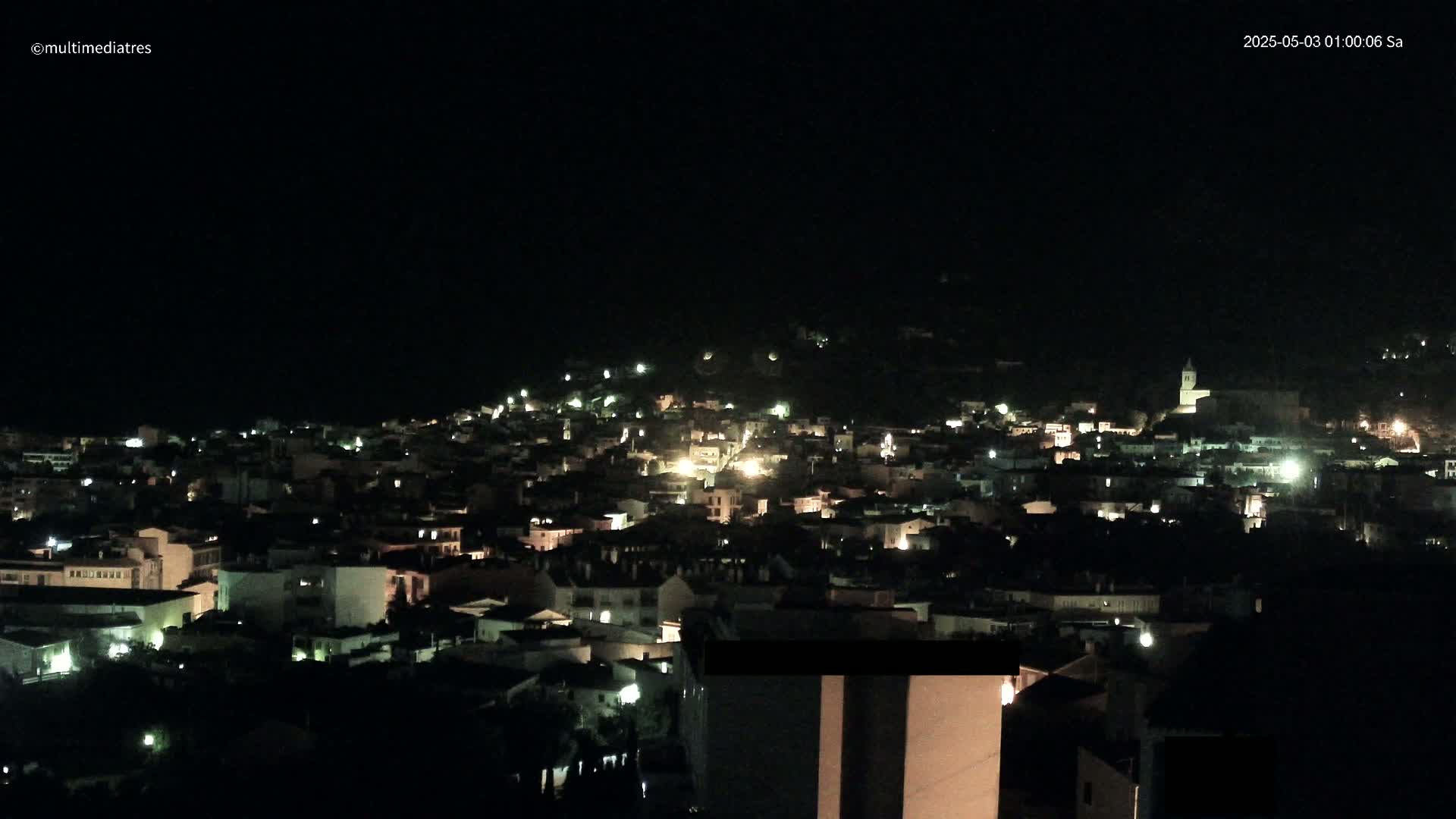 A nighttime, aerial view of a town nestled on a hillside, illuminated by scattered lights.