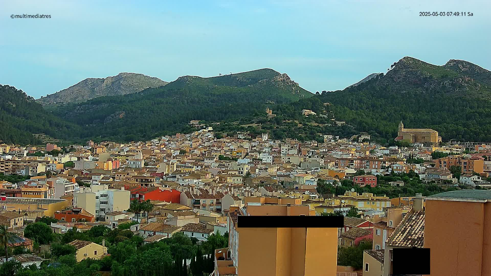 A sprawling town of low-rise buildings nestles at the foot of verdant, rocky hills under a clear, light blue sky.