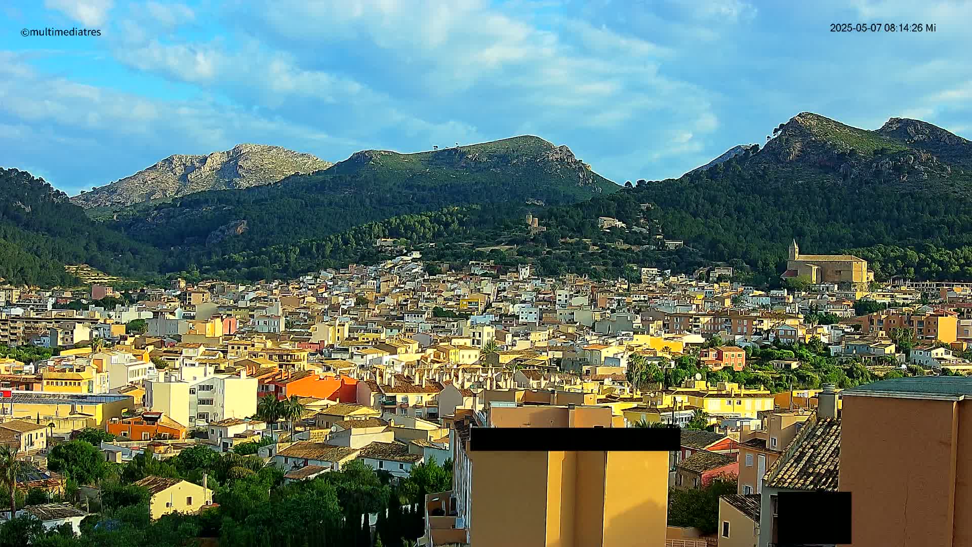 A sprawling town of predominantly yellow and orange buildings nestled at the base of a mountain range under a partly cloudy sky.