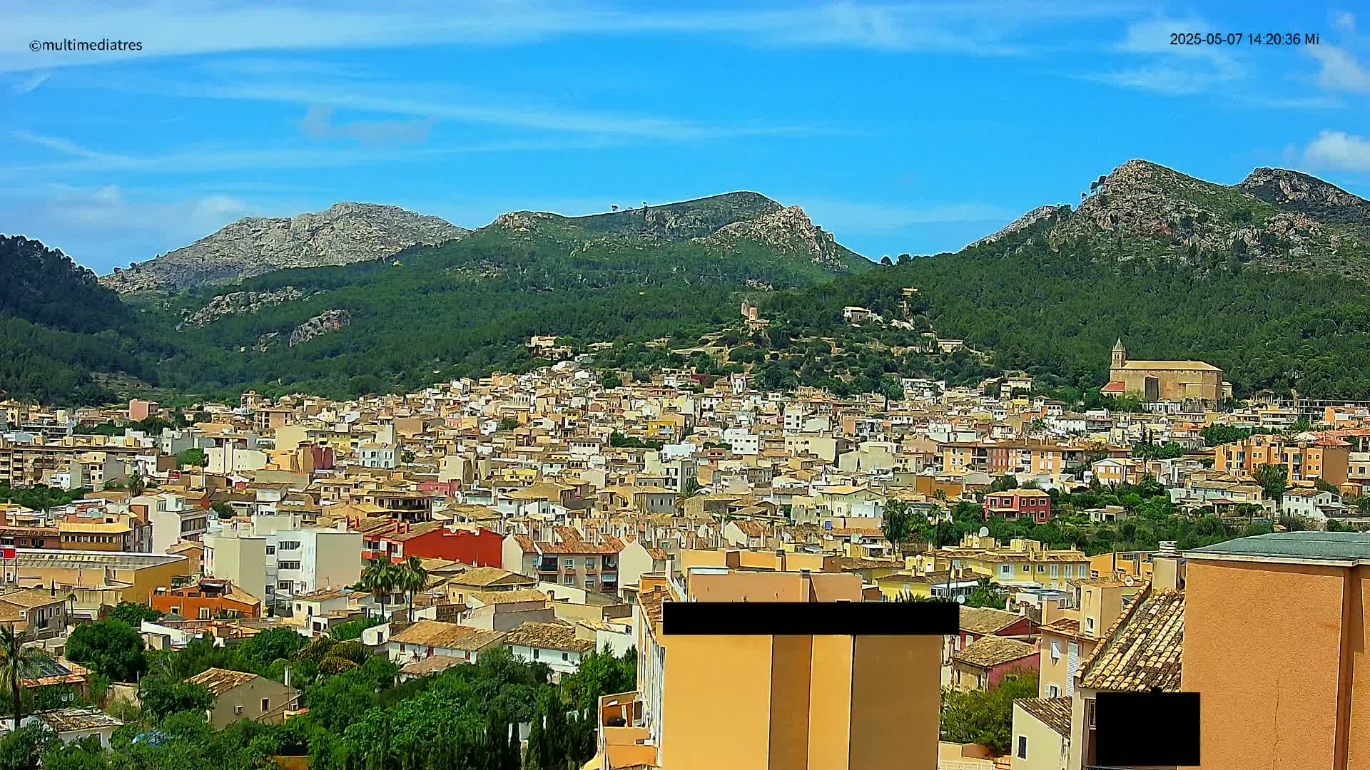 A dense collection of low-rise buildings extends from a hillside into a valley, nestled beneath mountains under a mostly clear, bright blue sky.