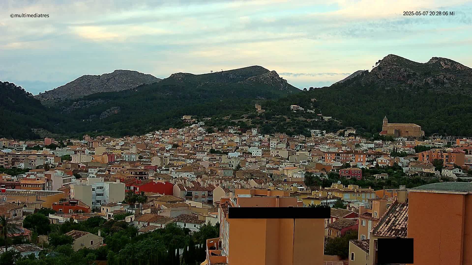 A sprawling town of low-rise buildings nestled at the base of tree-covered mountains under a partly cloudy sky.