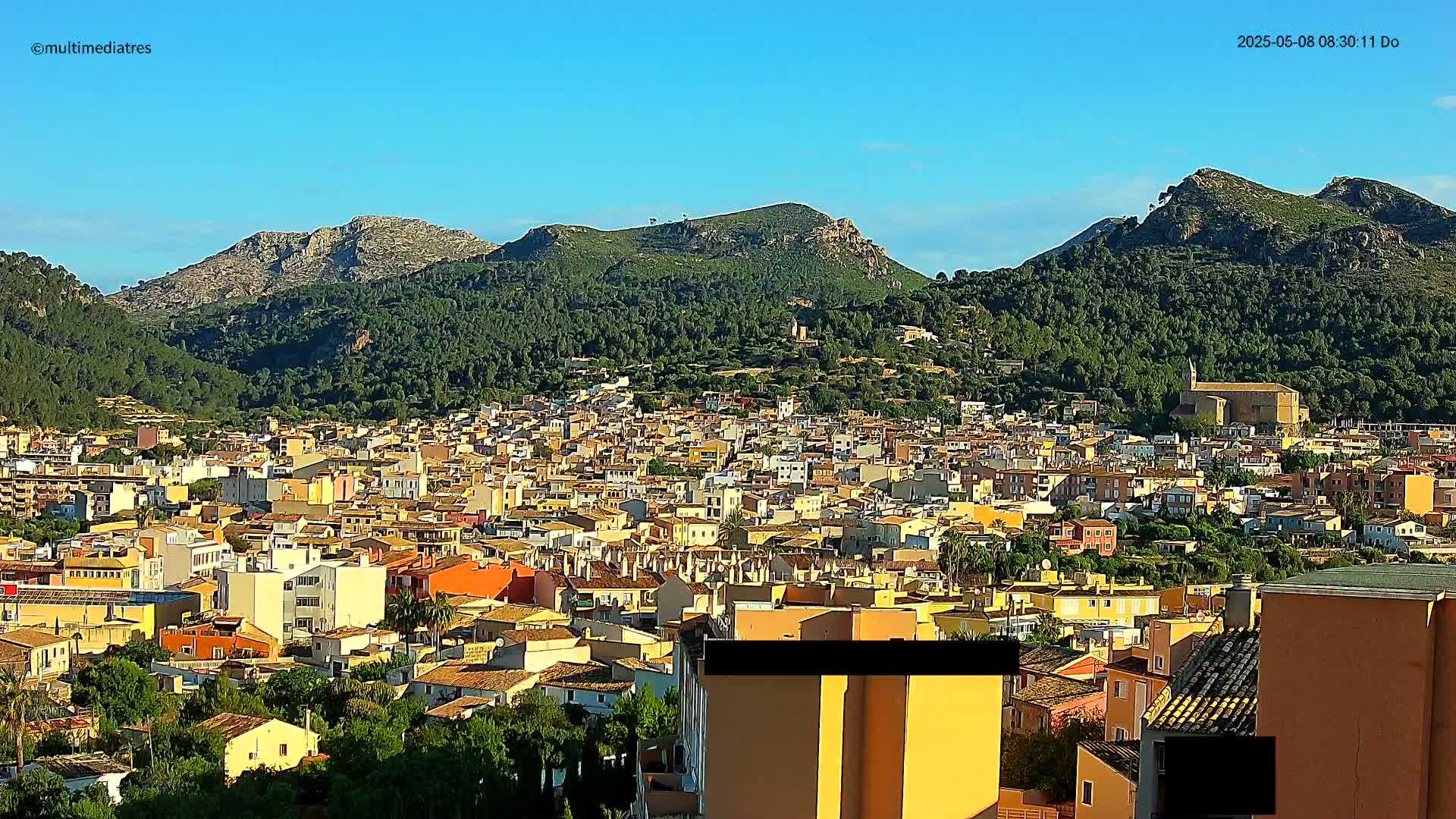 A sprawling town of closely packed, light-colored buildings nestles at the base of lush, green-covered mountains under a clear blue sky.