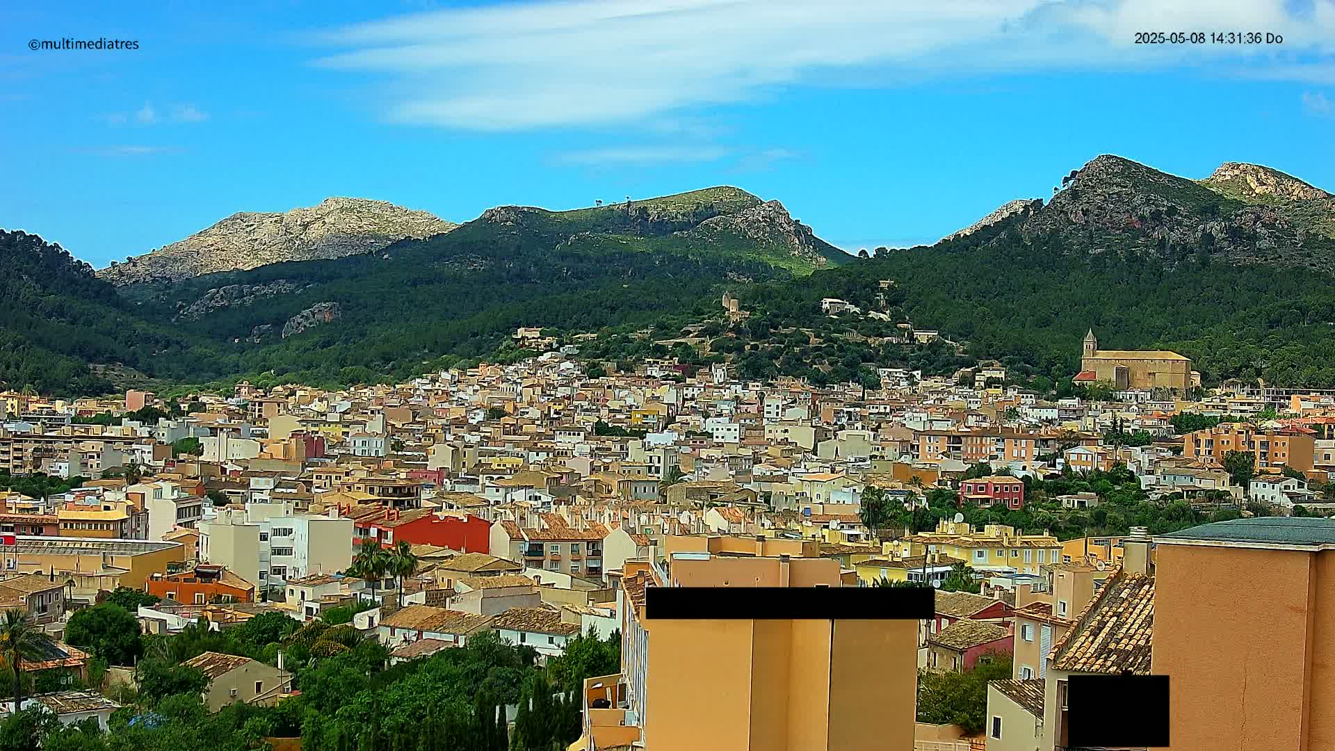 A sprawling town of tan and orange buildings nestles in a lush green valley surrounded by rocky mountains under a mostly clear blue sky.