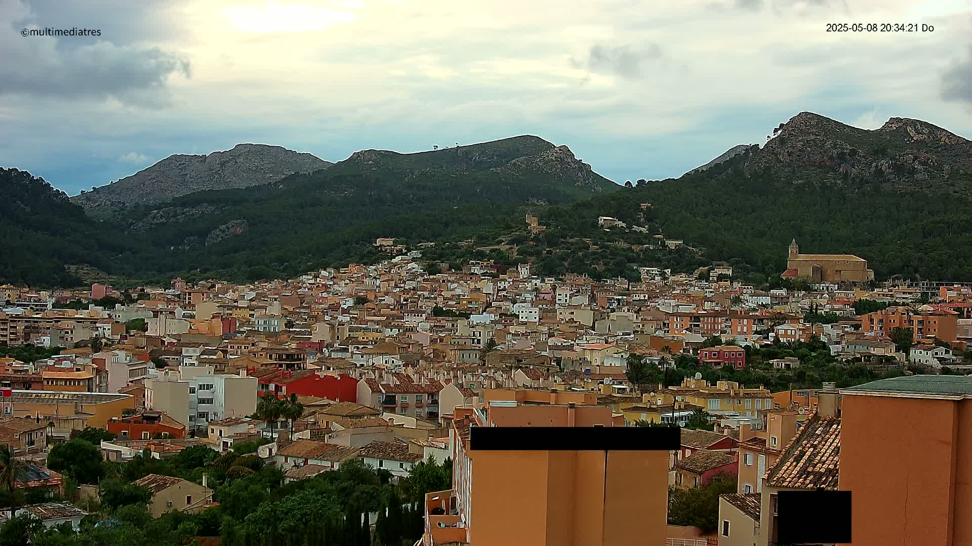 A sprawling town of low-rise buildings nestled in a valley between verdant, rocky mountains under a cloudy sky.