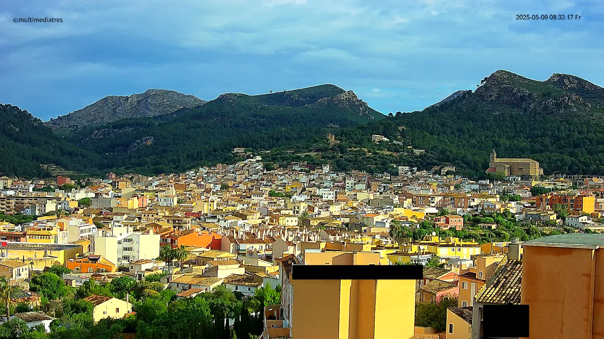 A dense collection of low-rise buildings sprawls across a hillside, backed by verdant mountains under a partly cloudy sky.