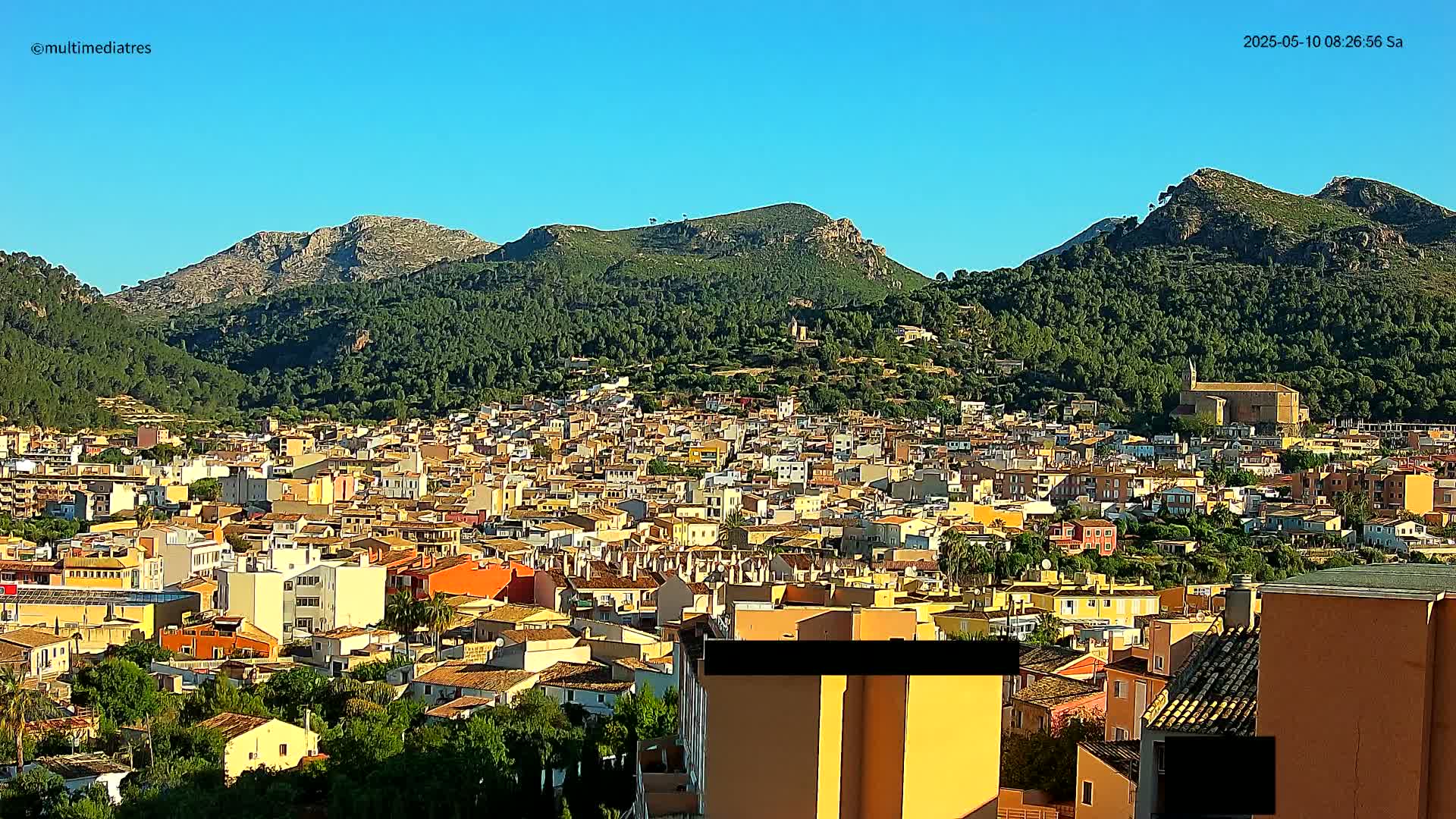 A dense cluster of low-rise buildings, predominantly yellow and orange, sprawls across a hillside, backed by lush green mountains under a clear blue sky.