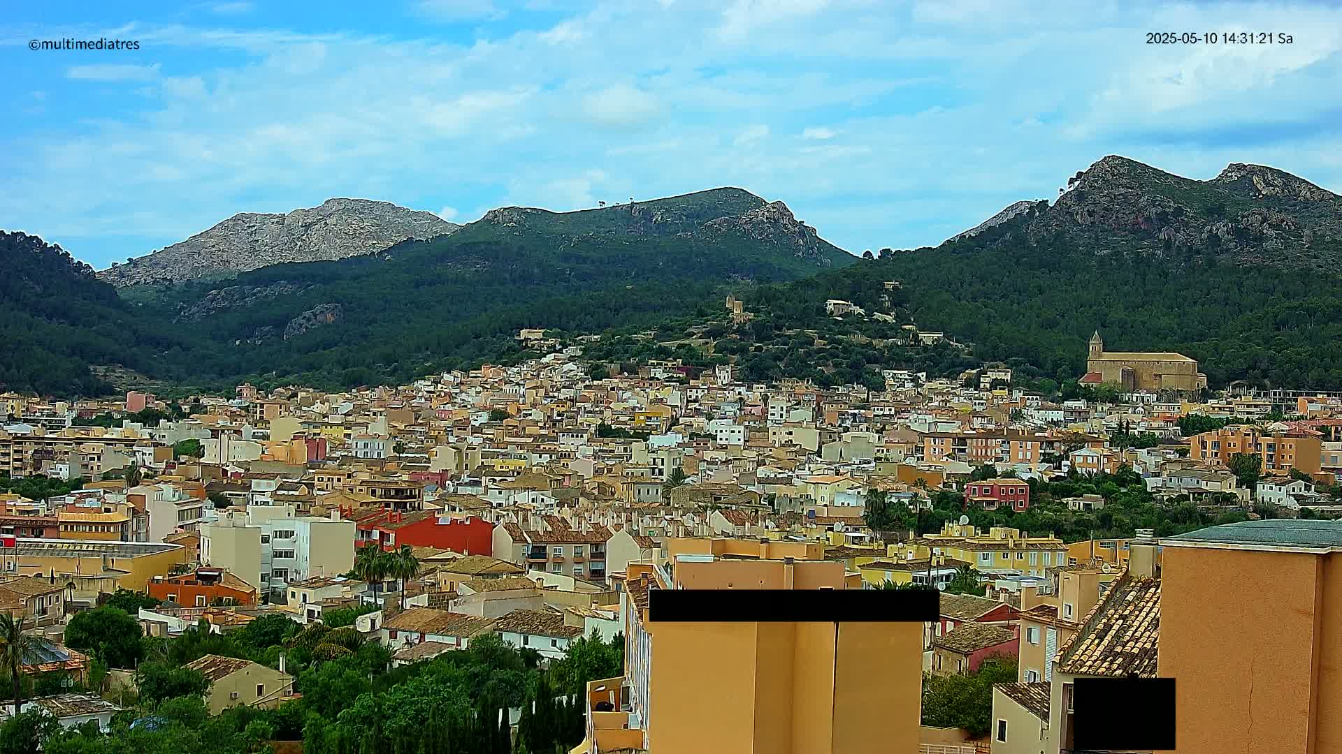 A sprawling town of colorful buildings nestled in a valley between verdant, rocky mountains under a mostly clear, bright blue sky.