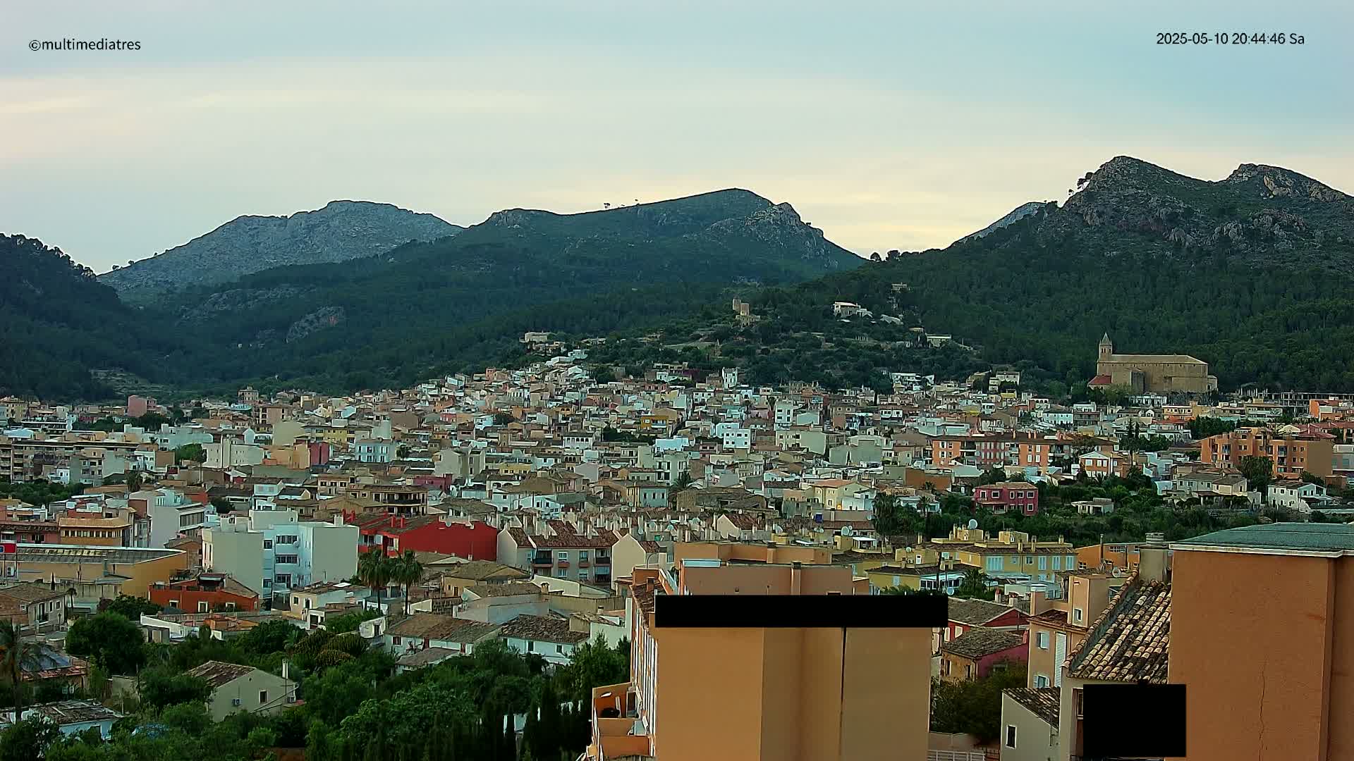 A dense collection of buildings nestled in a valley, surrounded by green hills and mountains under a cloudy sky.