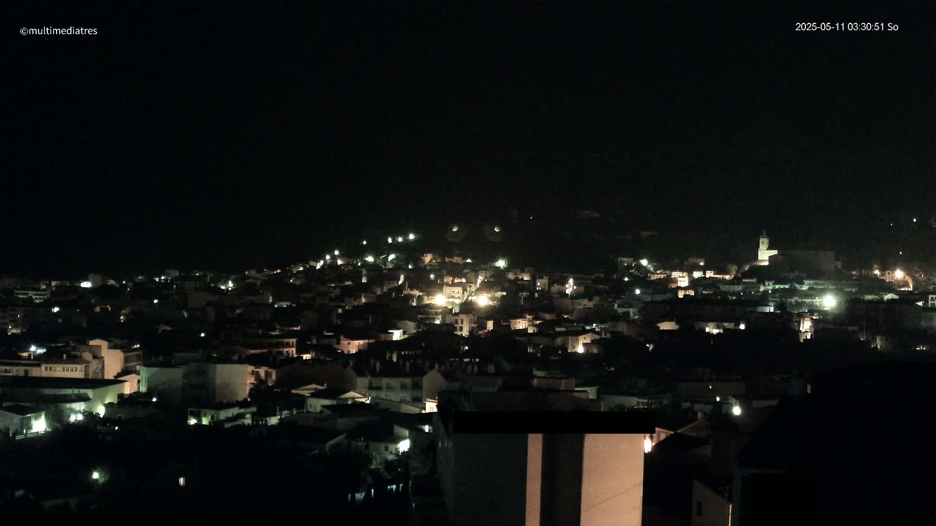 A nighttime view of a town nestled on a hill, with scattered lights illuminating the buildings and streets under a dark sky.